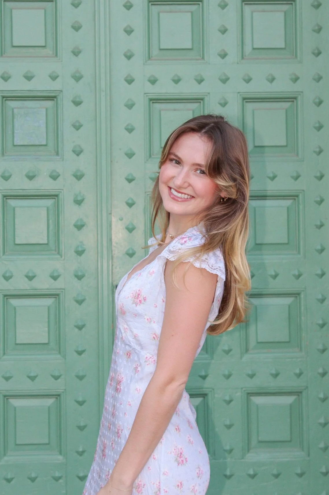 A young woman with long, wavy hair smiling in front of a green door with decorative square and diamond patterns.