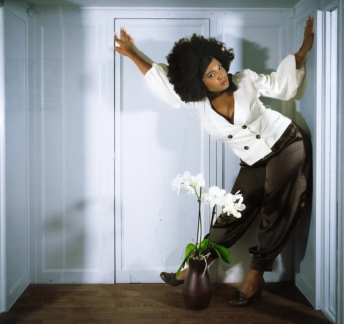 A woman with curly hair wearing a white blazer and dark pants strikes a pose inside a small corner with white walls, with a brown vase holding white orchids at her feet.