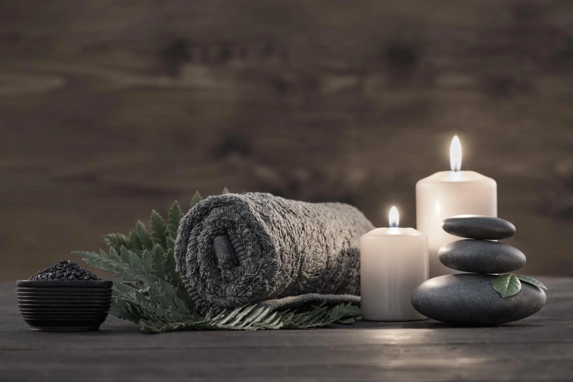 A neatly rolled gray towel, a small black bowl with black grains or seeds, green leaves, two lit white candles, and a stack of three smooth black stones with green leaves, all arranged on a wooden surface for a spa or relaxation setting.