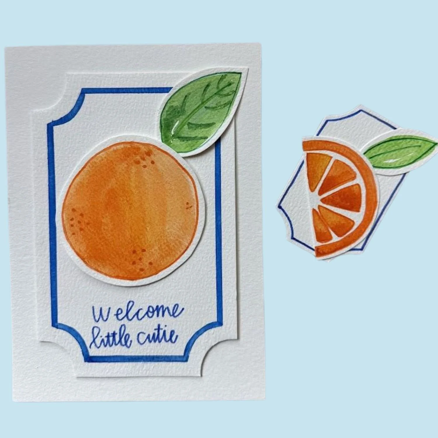 Watercolor-style orange with a green leaf on a white card that says 'Welcome little cutie,' next to a watercolor orange slice with a leaf on a light blue background.