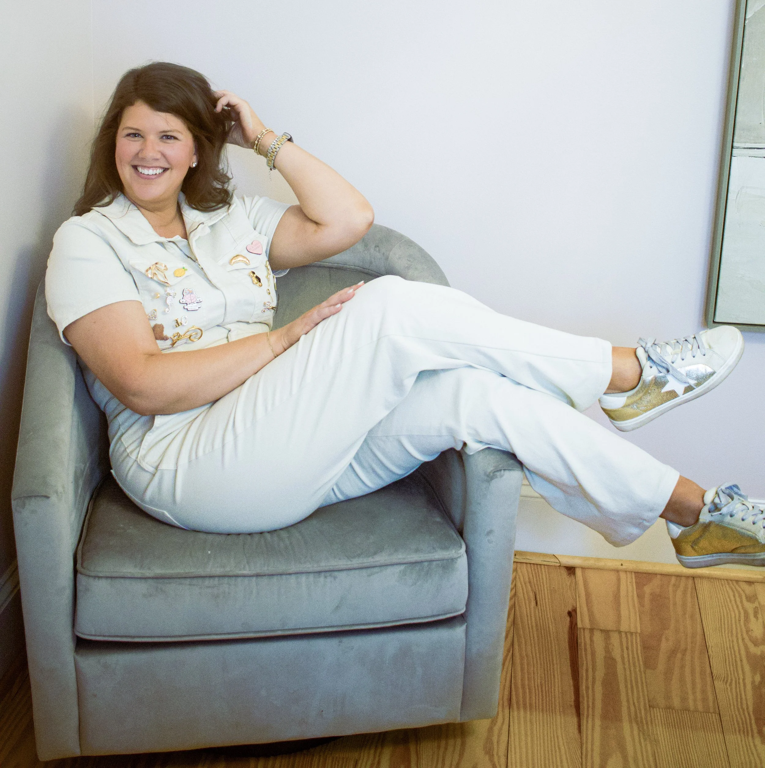 A woman with brown hair, smiling, sitting on a gray armchair with her legs crossed at the knee and feet resting on the floor, wearing a light-colored uniform with patches and gold sneakers.