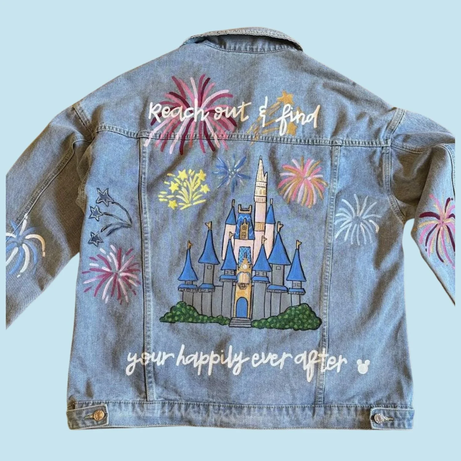 Back of a custom painted denim jacket with a colorful Disney castle and fireworks painted on it, along with the text 'reach out & find your happily ever after'.