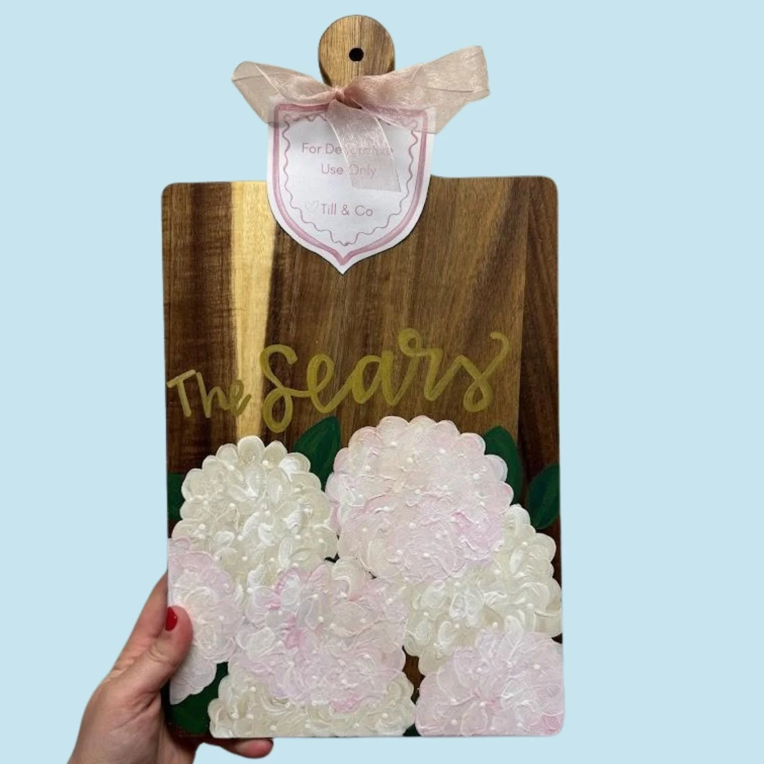 Hand holding a decorative cutting board with pink hydrangea flowers and the words 'The Seas' written in gold, against a plain light blue background.