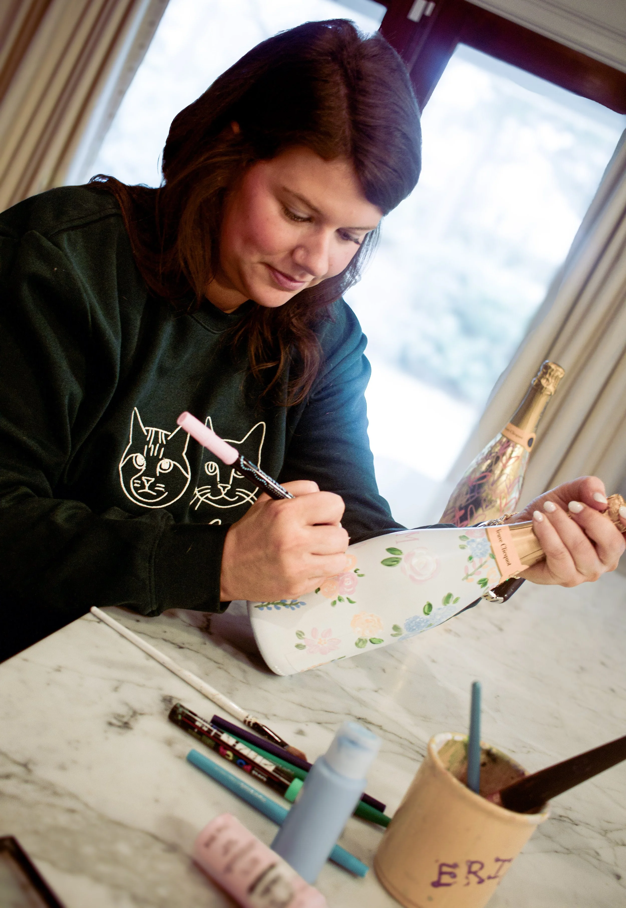 Woman customizing a floral-patterned champagne bottle with a pink marker.