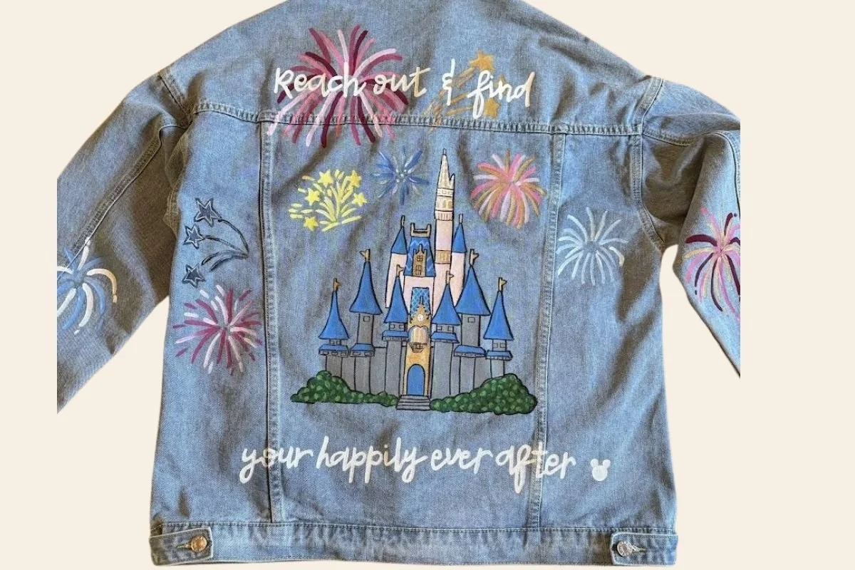Back of a denim jacket decorated with colorful fireworks, a castle, and the phrases "reach out & find" and "your happily ever after".
