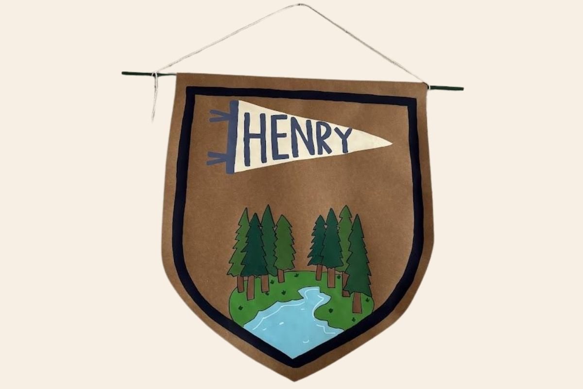 A fabric wall hanging with a brown background featuring a forest scene with green pine trees and a river, and a white pennant with the name "Henry" in blue letters.