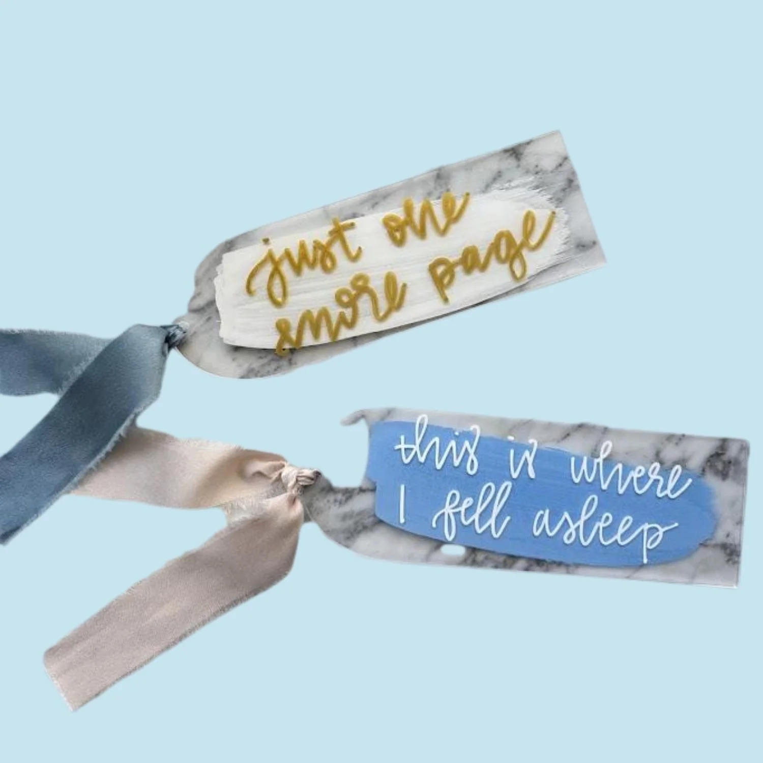Decorative fabric pieces with inspiring messages on marble-patterned paper, attached to ribbons. One says 'just one more page,' and the other says 'this is where I fell asleep.'