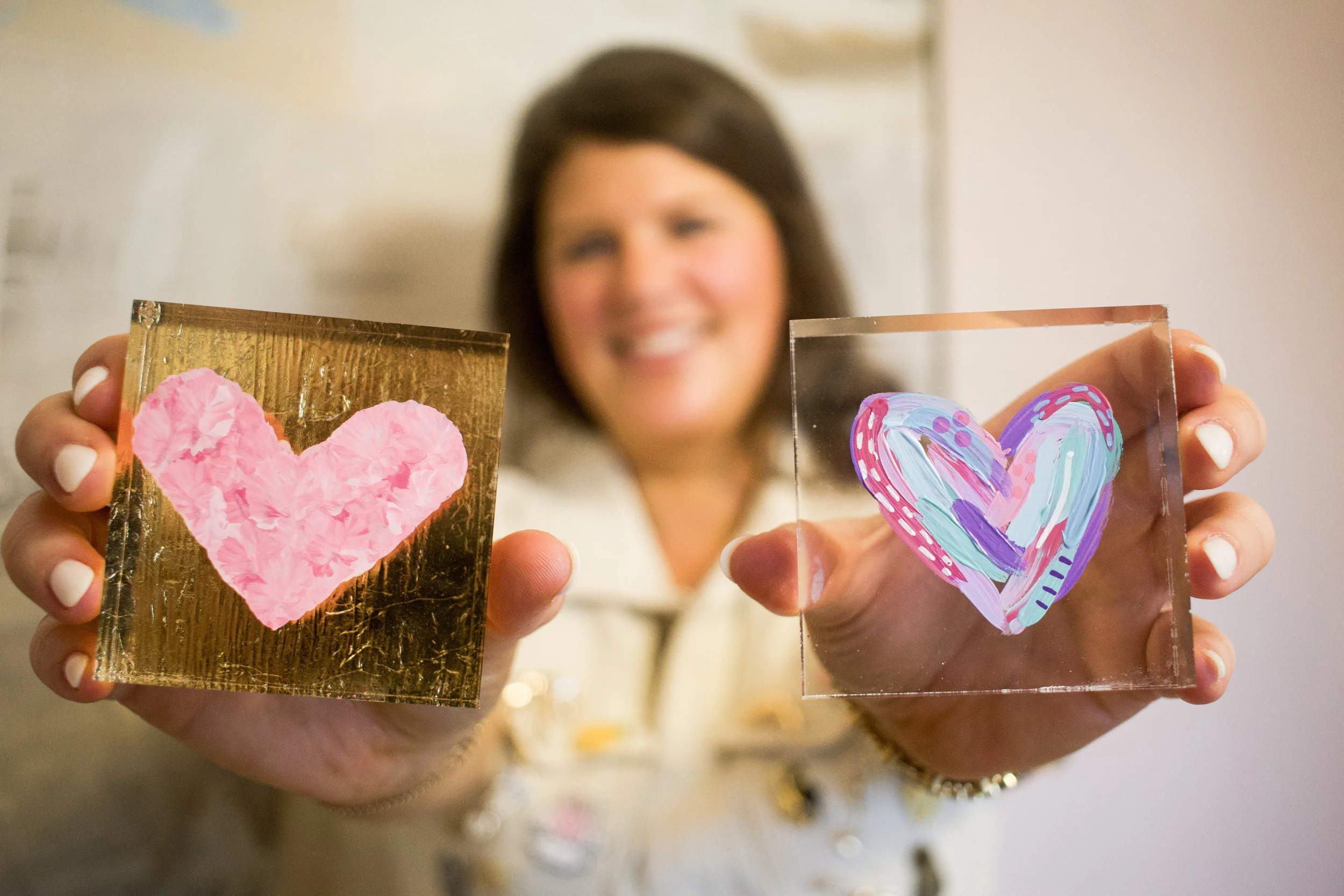 A woman holding up two pieces of artwork, each with a colorful heart design. The left heart is pink on a gold background, and the right heart is multicolored on a transparent square.