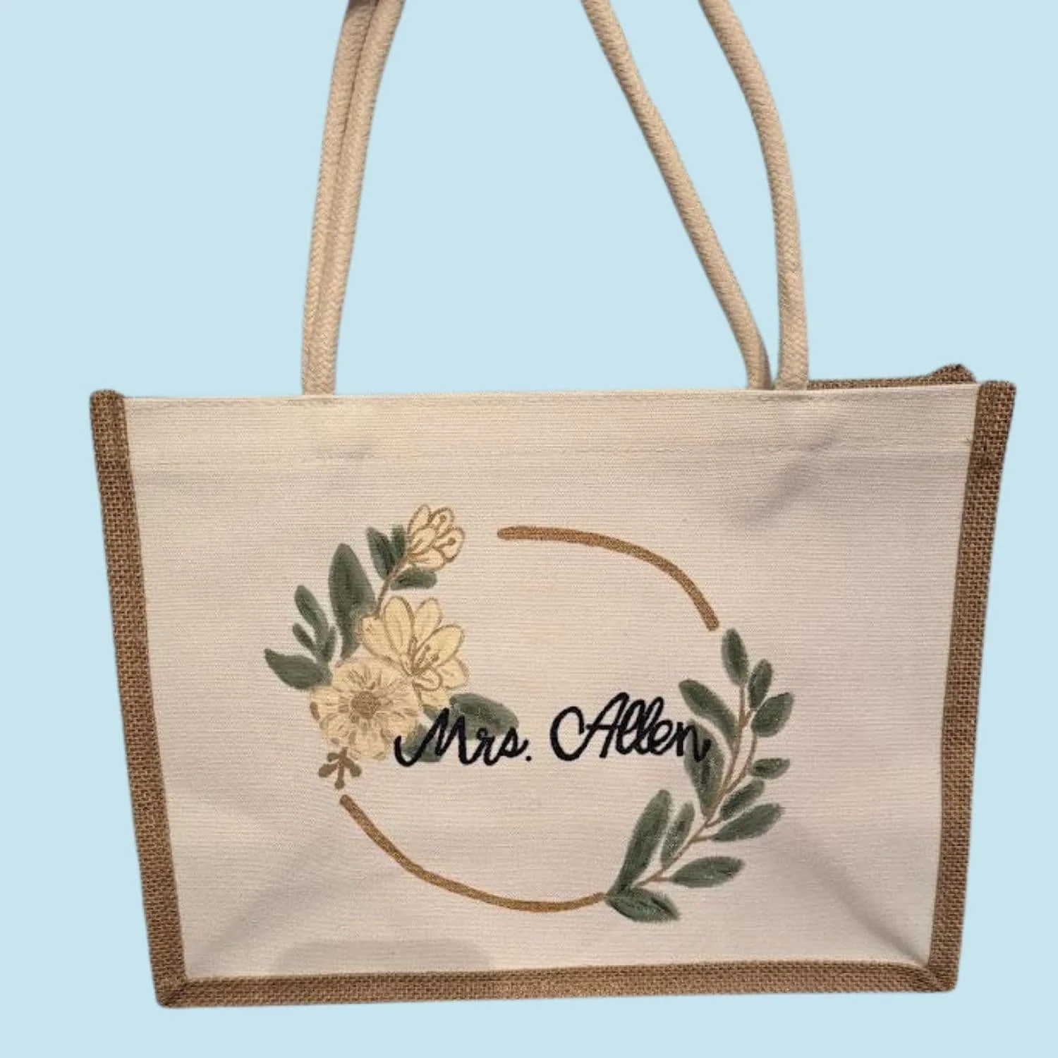 Beige canvas customized tote bag with brown edges and handles, featuring a floral wreath and the text 'Mrs. Allen' in black script on the front.