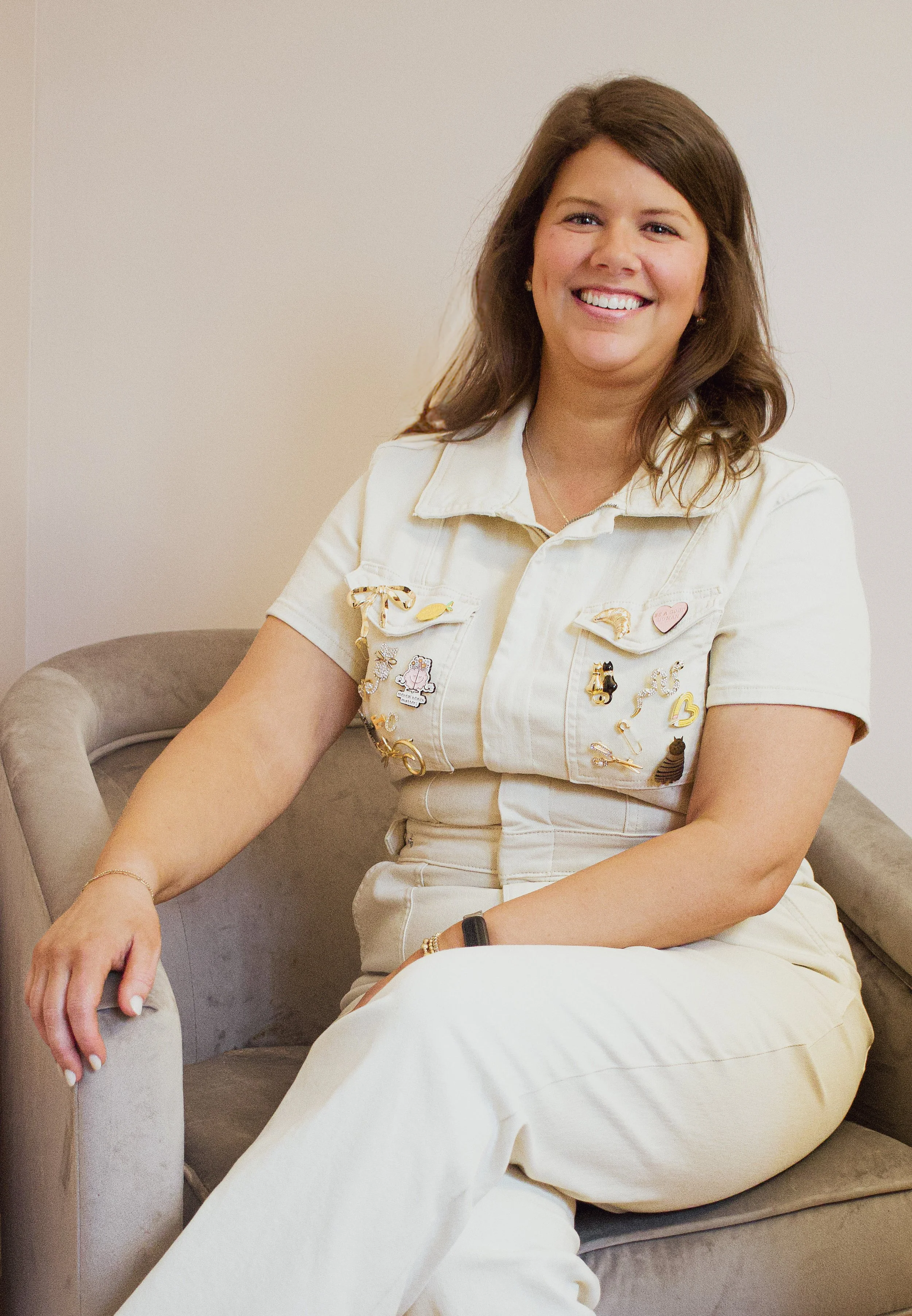 A woman with brown hair smiling and sitting on a beige sofa, wearing a cream-colored outfit adorned with various pins.