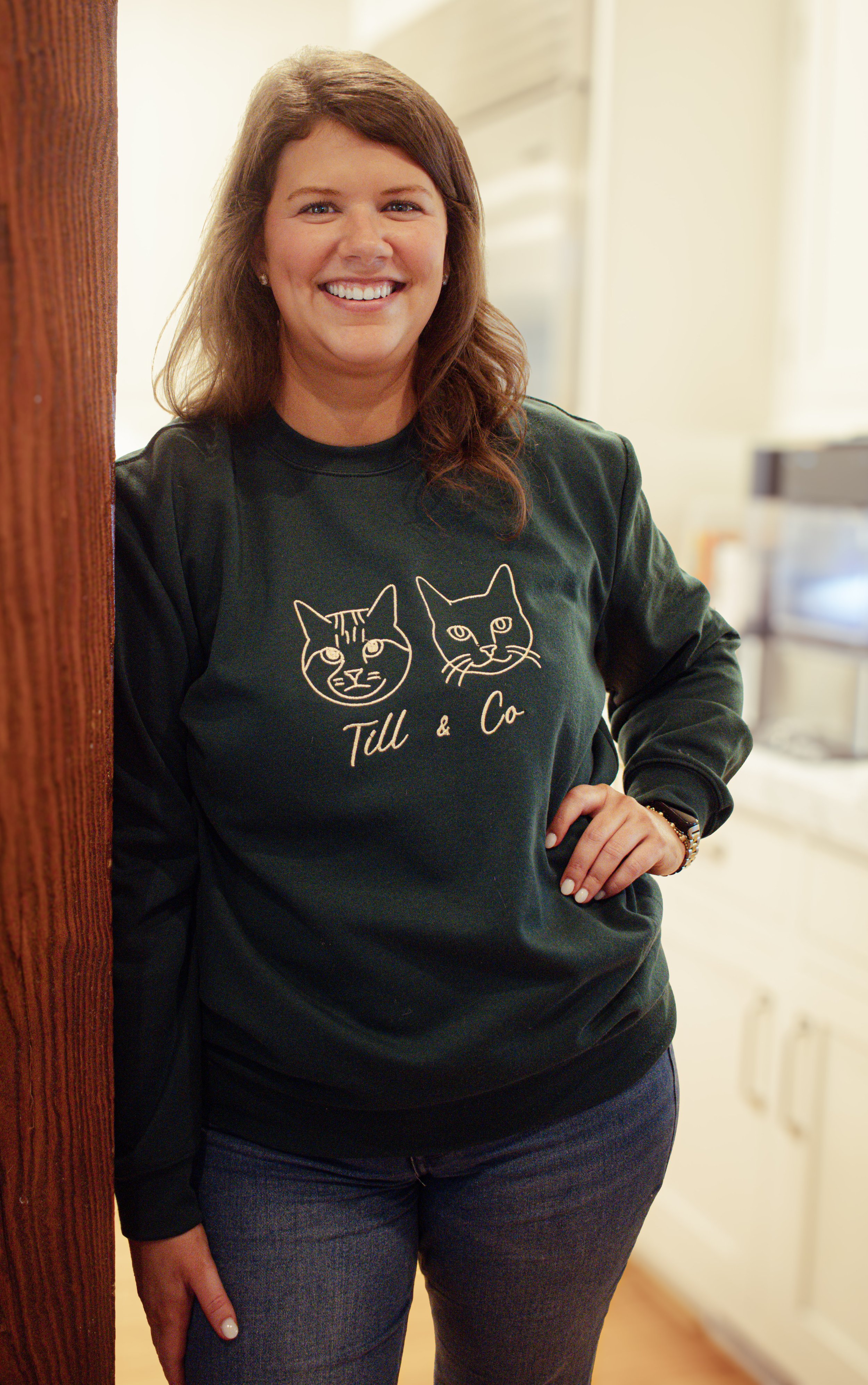 A woman with long brown hair smiling, wearing a black sweatshirt with embroidered outline of two cats and text 'Till & Co' standing in a room with a wooden door frame and light-colored kitchen in the background.
