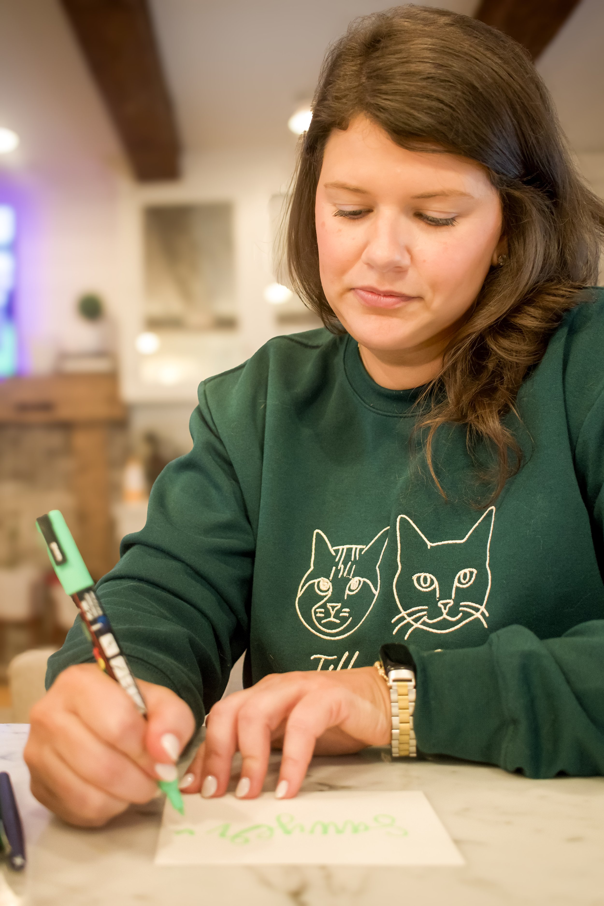 A woman with brown hair wearing a green sweatshirt with two cat faces on it is writing with a colorful marker on a piece of paper.
