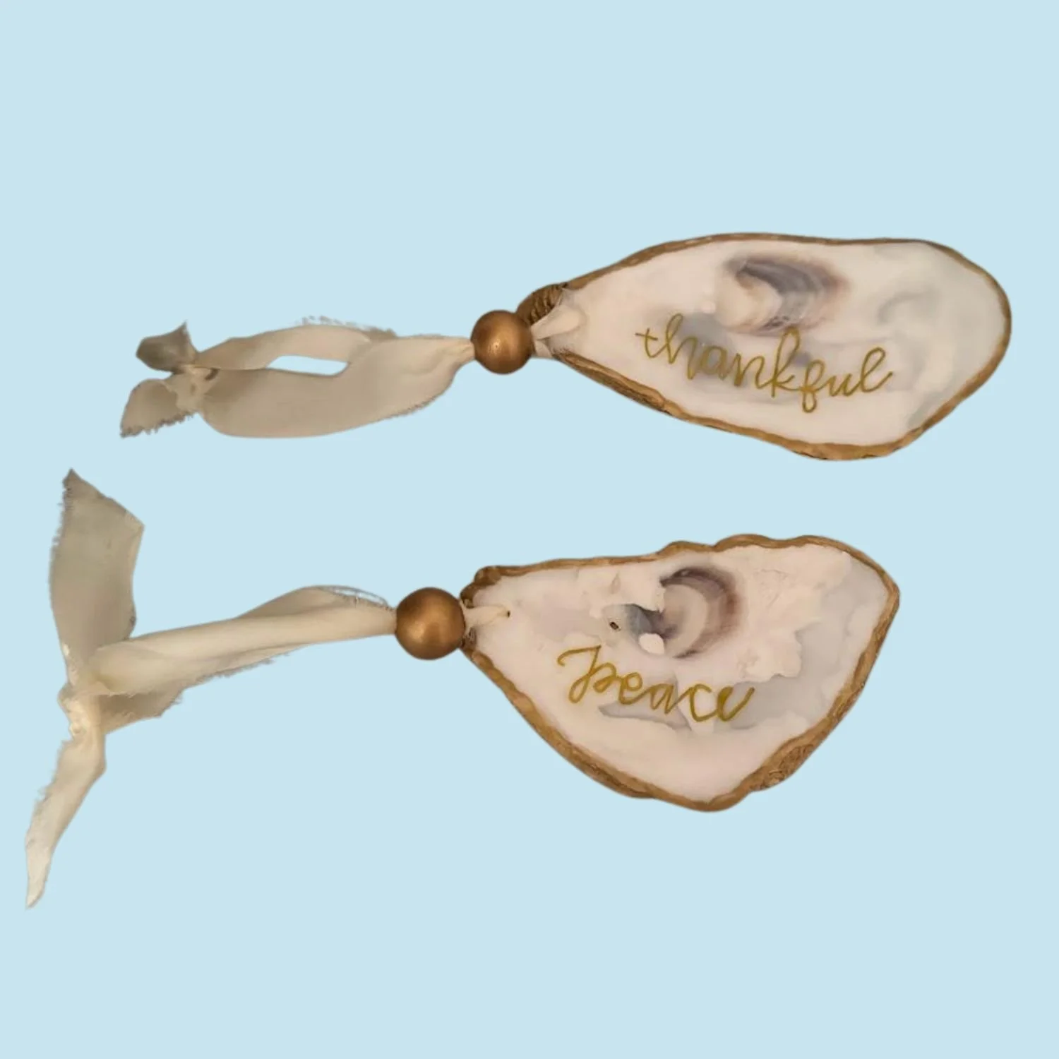 Two custom decorative oyster shell ornaments with gold dot accents and beige ribbons, each inscribed with words 'thankful' and 'peace' in gold script, on a light blue background.