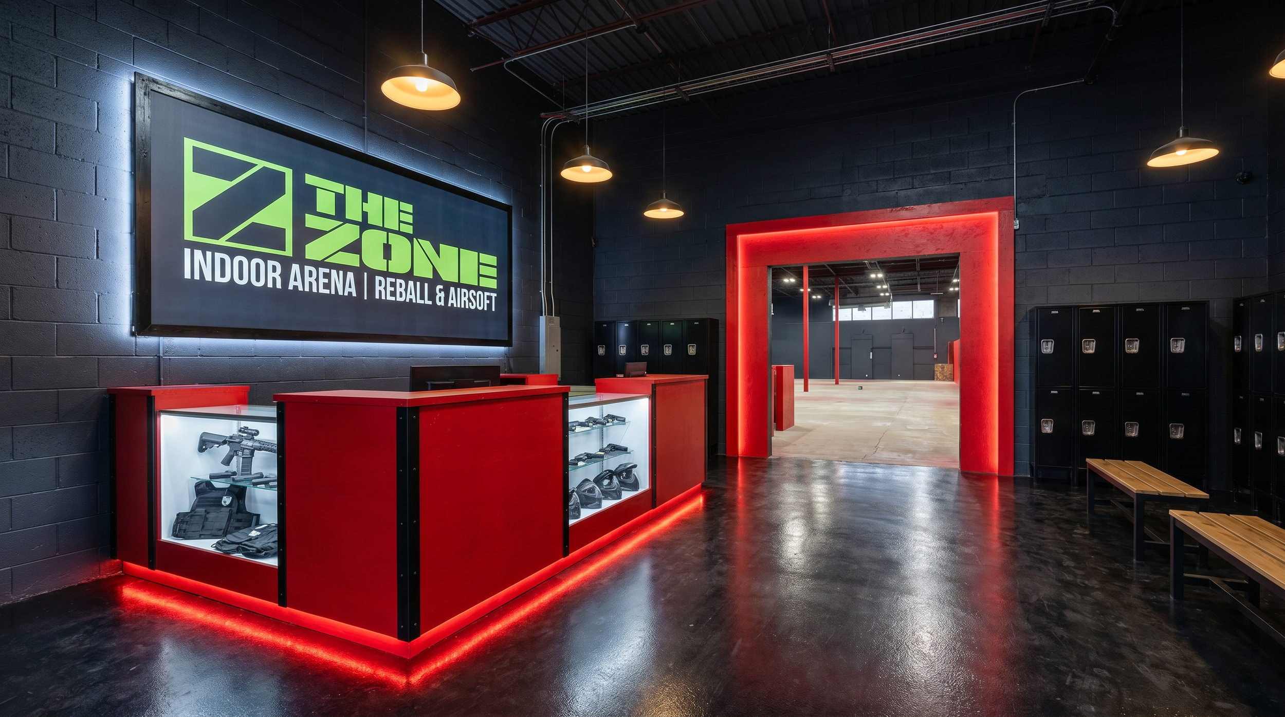 Interior of an indoor arena with black brick walls, a red-lit entrance, lockers, a display case with paintball gear, and a large digital sign for 'The Zone Indoor Arena Reball & Airsoft.'