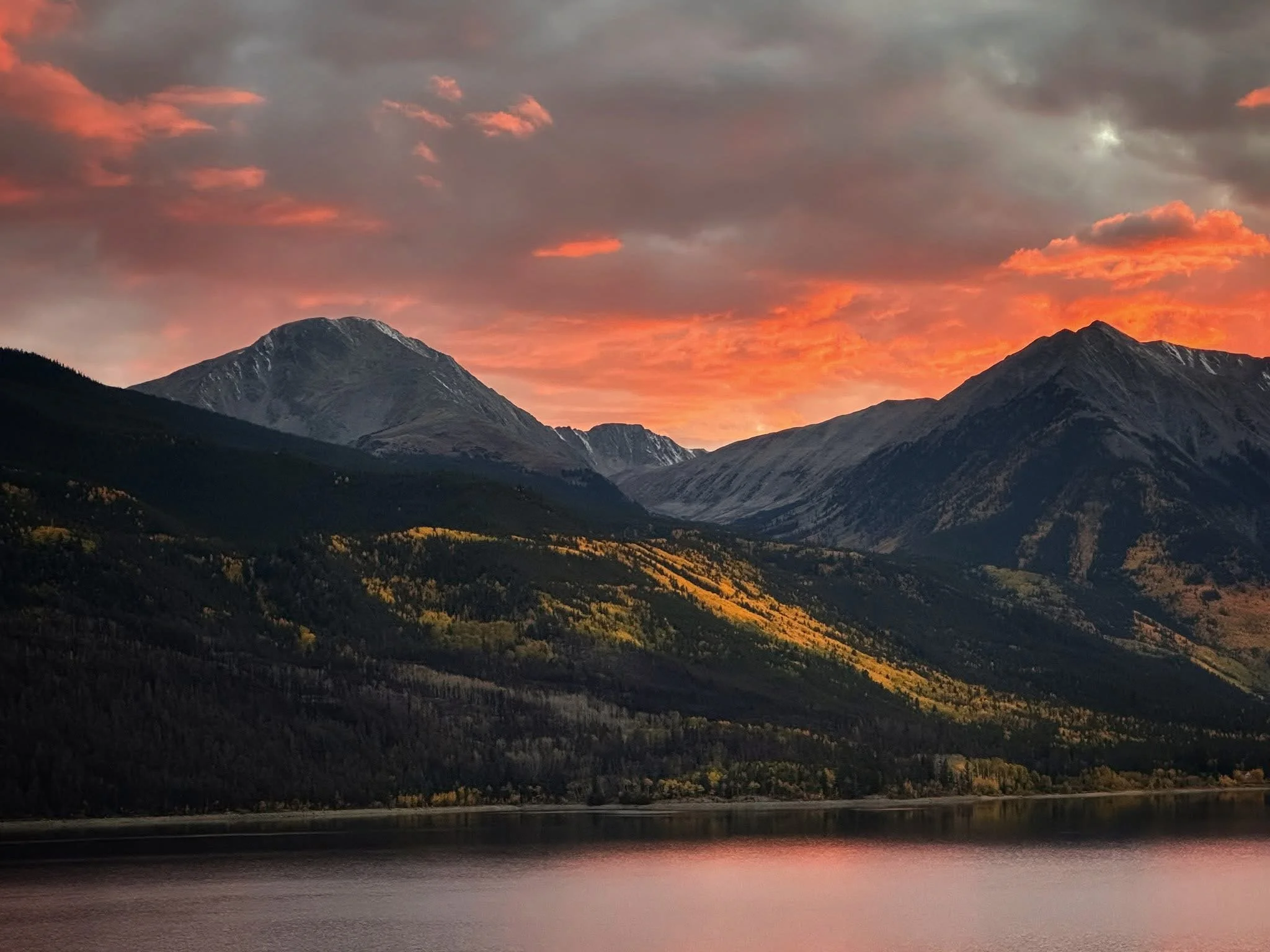 A mountain landscape at sunset with a sky filled with pink and orange clouds, dark mountains, a forested area, and a body of water in the foreground.