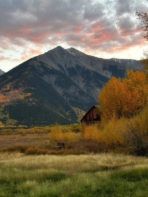 A mountain landscape with a mountain peak, colorful autumn trees, a rustic wooden barn, and a grassy field at sunset.