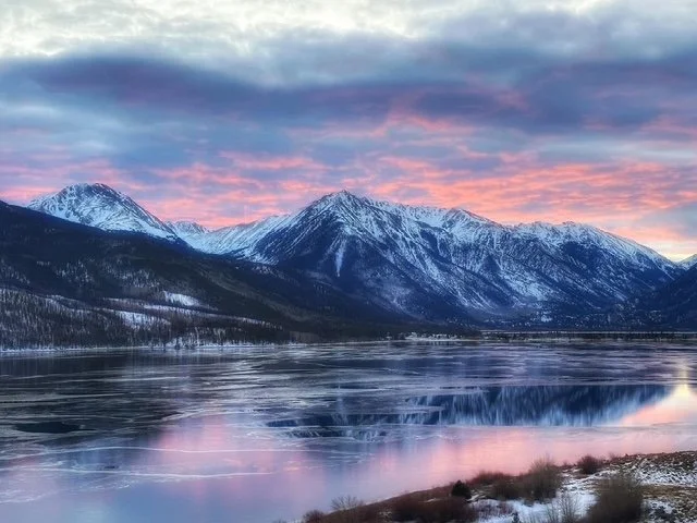 Snow-capped mountains reflect in a partially frozen lake during a colorful sunset.