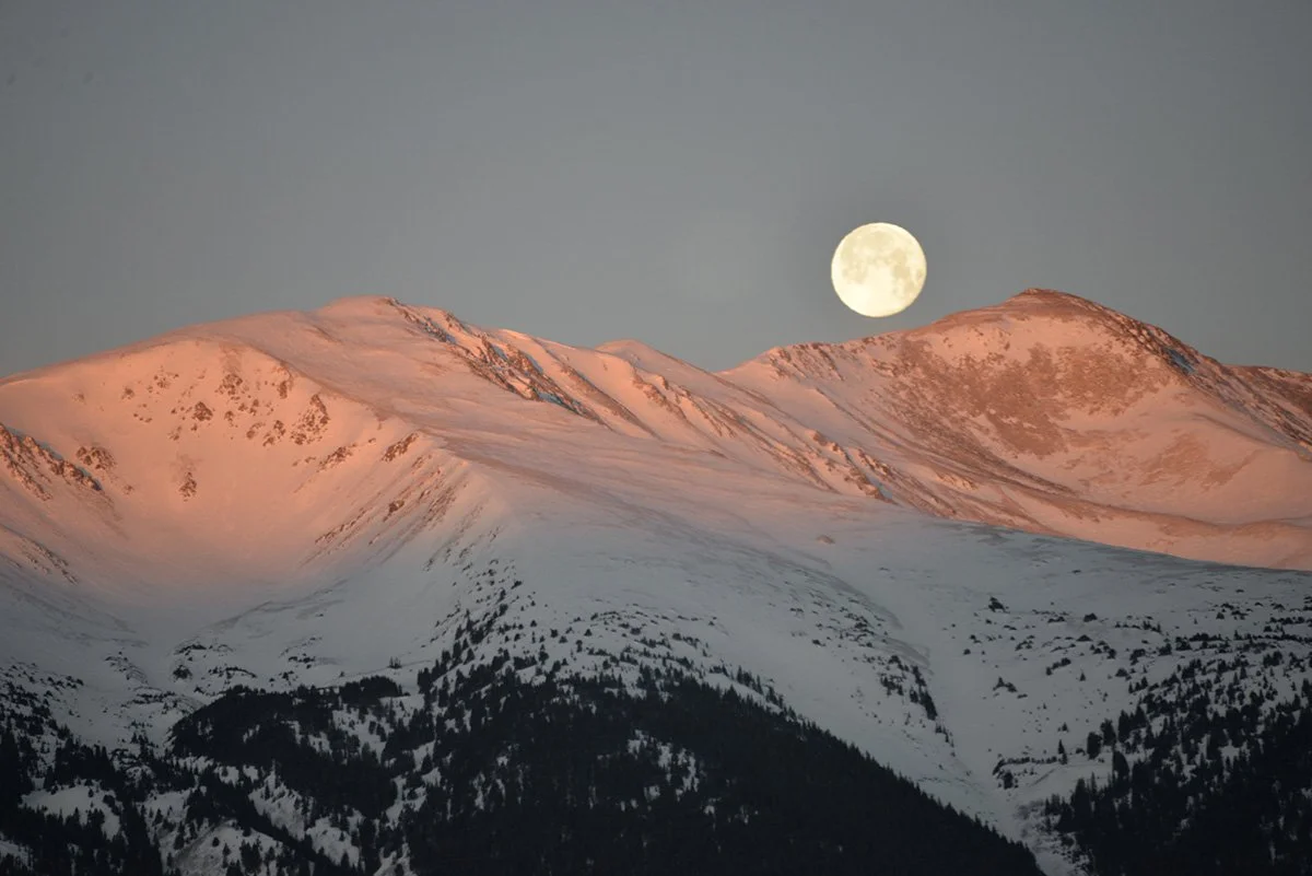 Snow-covered mountain range under a full moon with a clear sky.