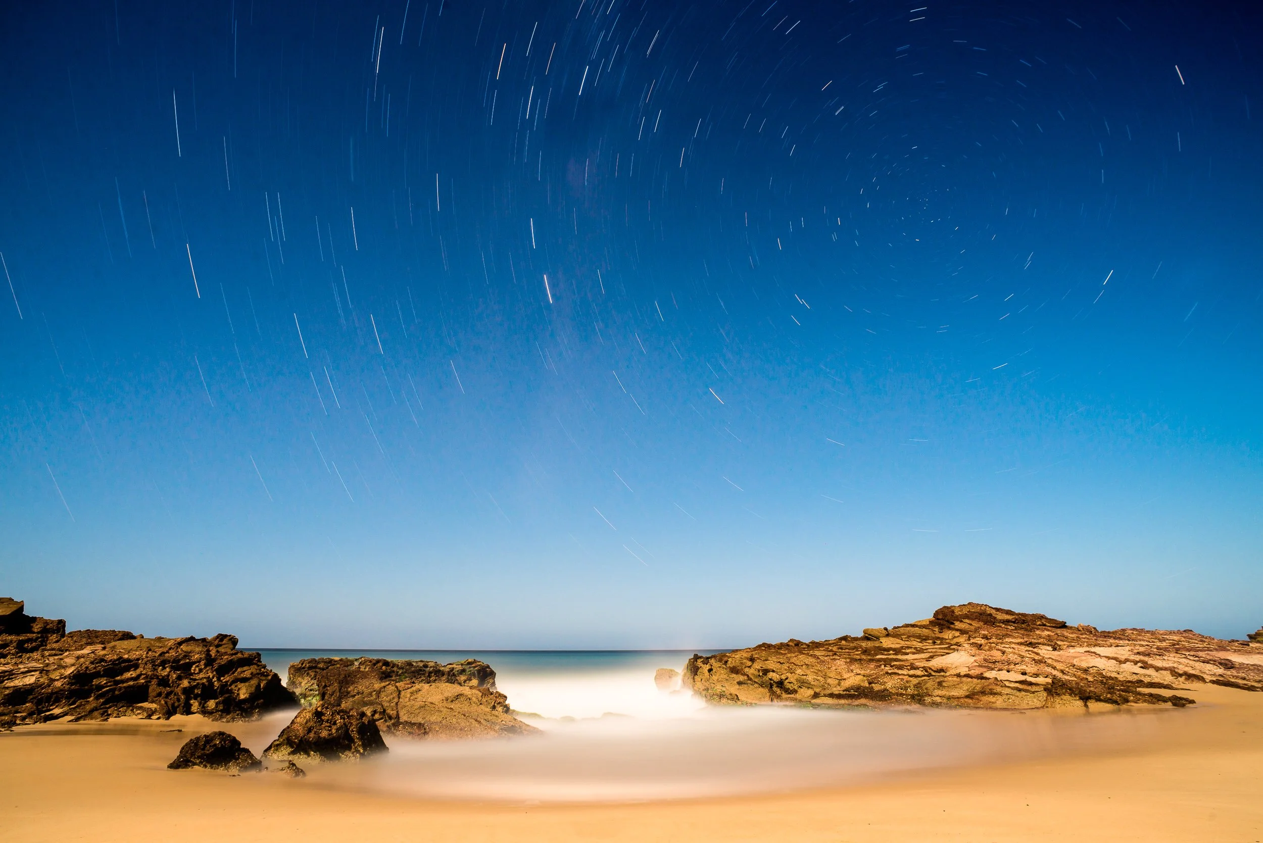 Nighttime beach scene with star trails in the sky, rocky shoreline, and sandy beach in the foreground.