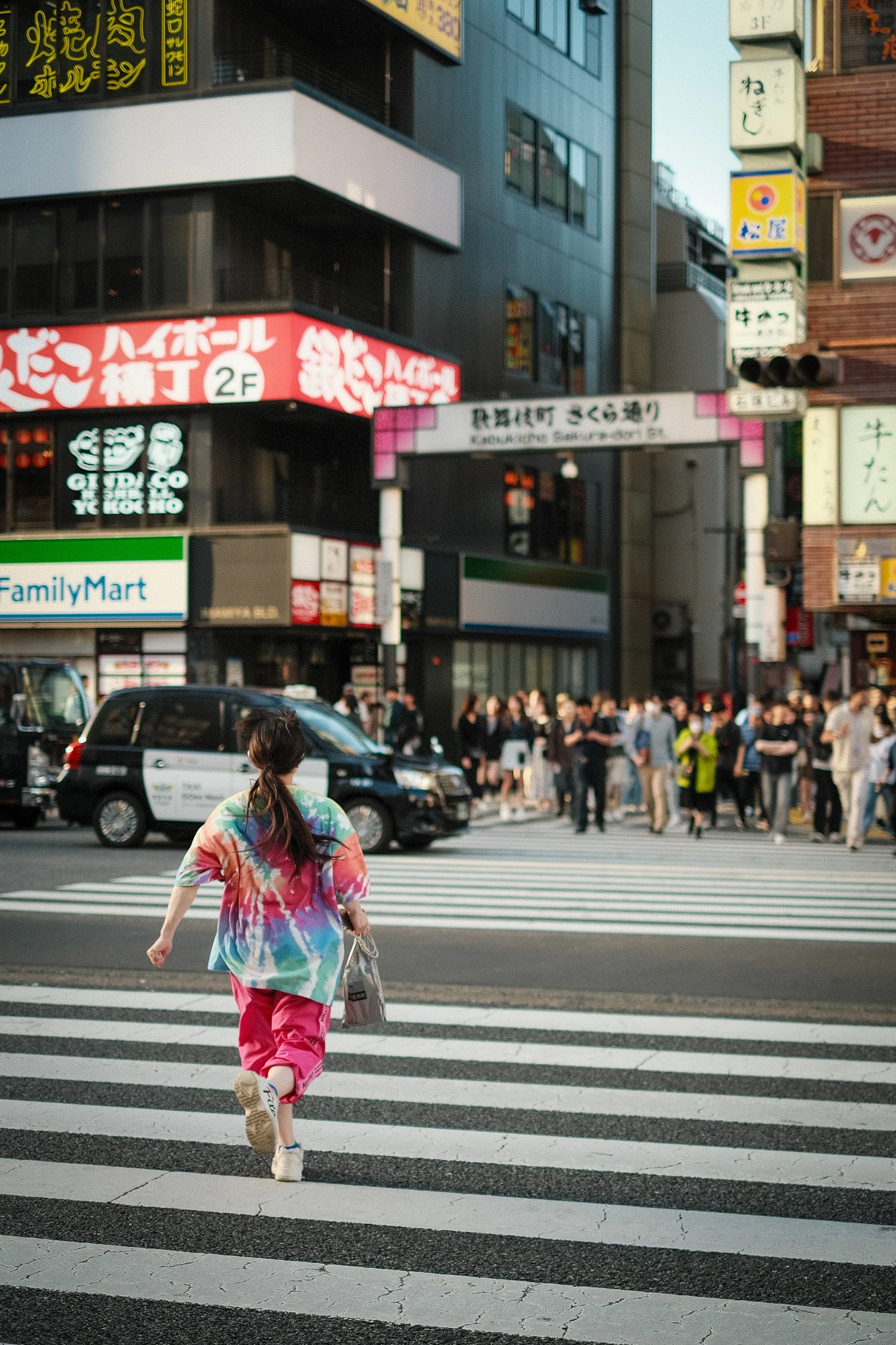 A woman in a colorful tie-dye shirt and pink pants crossing a street at a busy city intersection with many people and tall buildings with illuminated signs in the background.