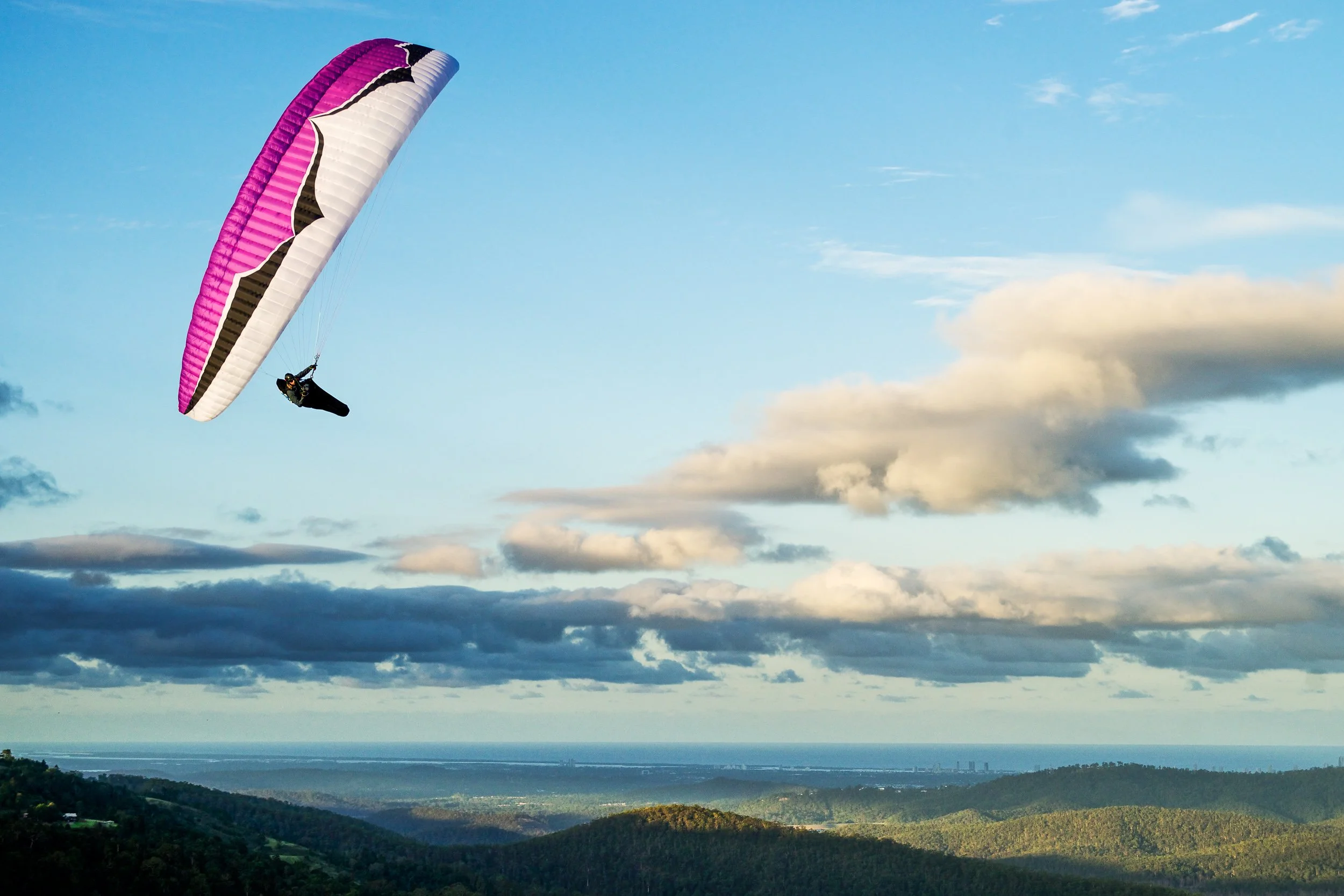 A person paragliding with a pink and white parachute over a landscape of hills and forests, with a cloudy sky above.