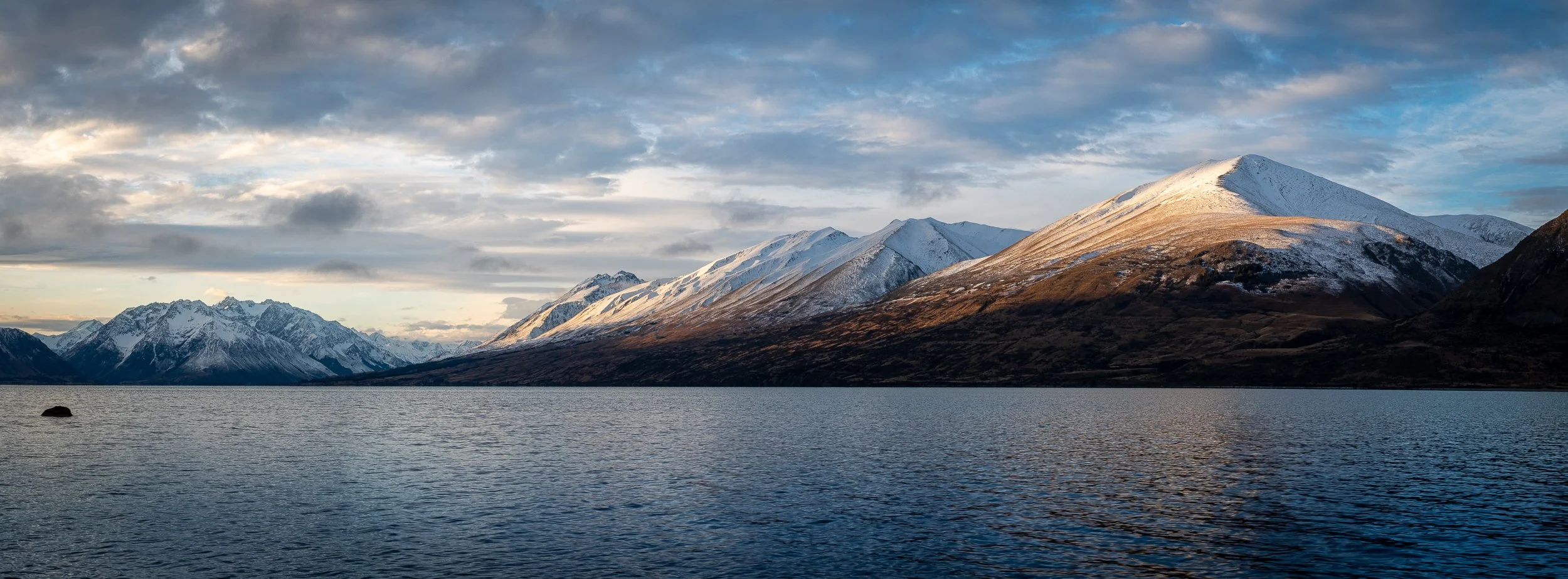 A serene lake surrounded by snowy mountains under a partly cloudy sky at sunset.