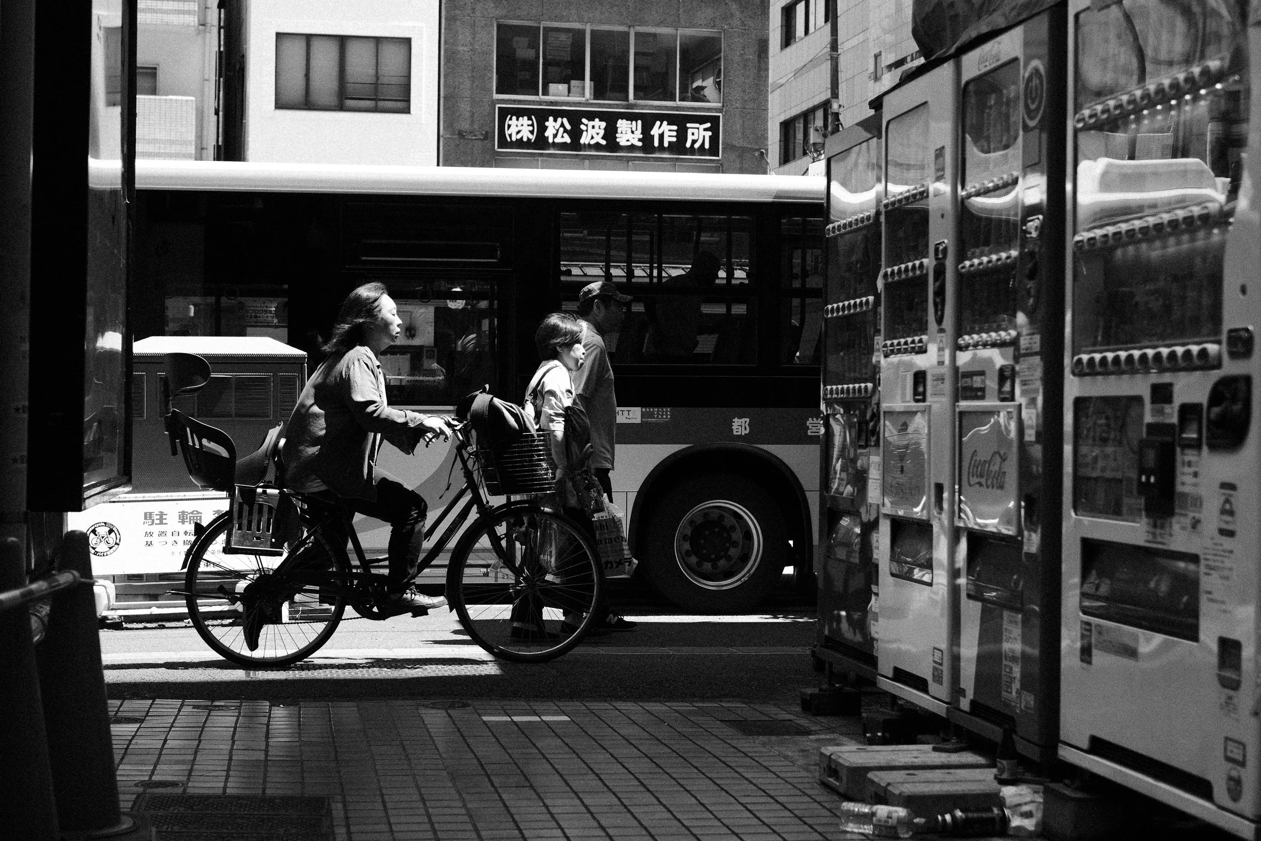 A street scene in an urban area with a woman riding a bicycle, a person walking, and a bus passing by. There are vending machines on the right and buildings in the background with store signs.