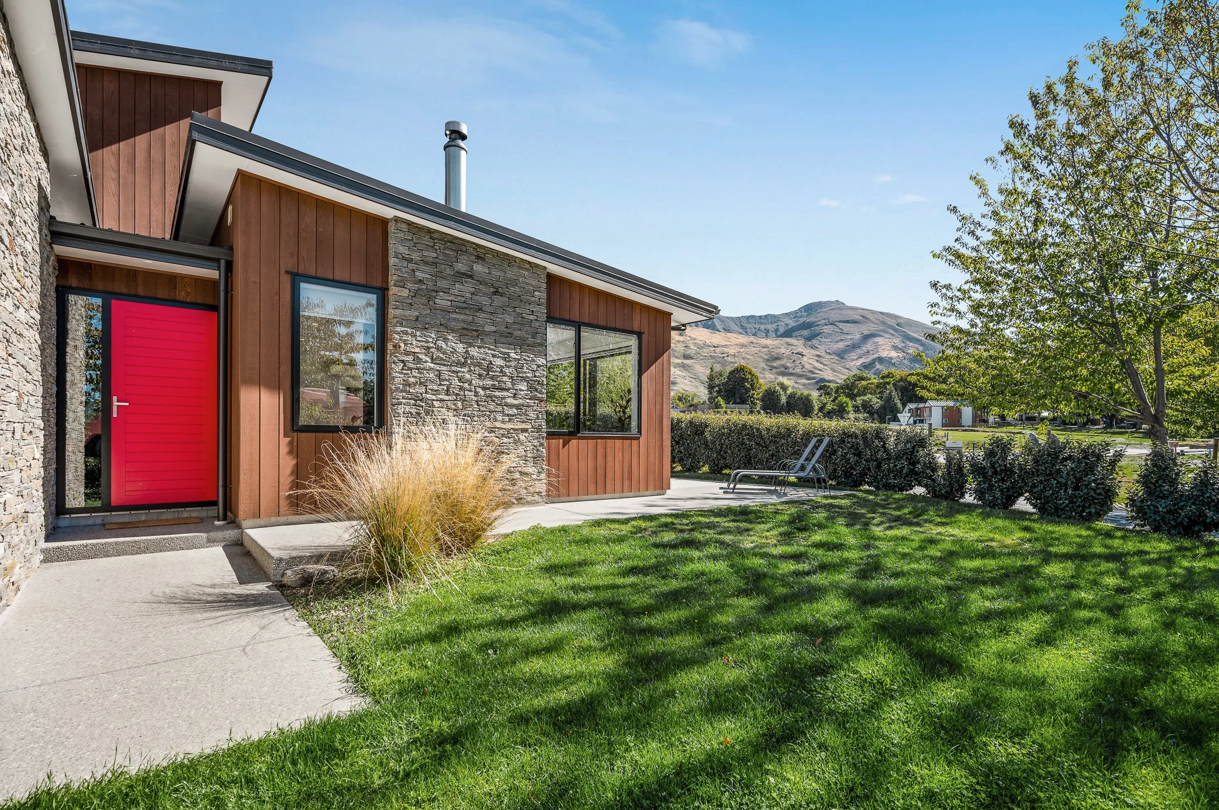 Modern house with a red door, large windows, a stone and wood exterior, and a well-maintained lawn with trees and mountain view in the background.