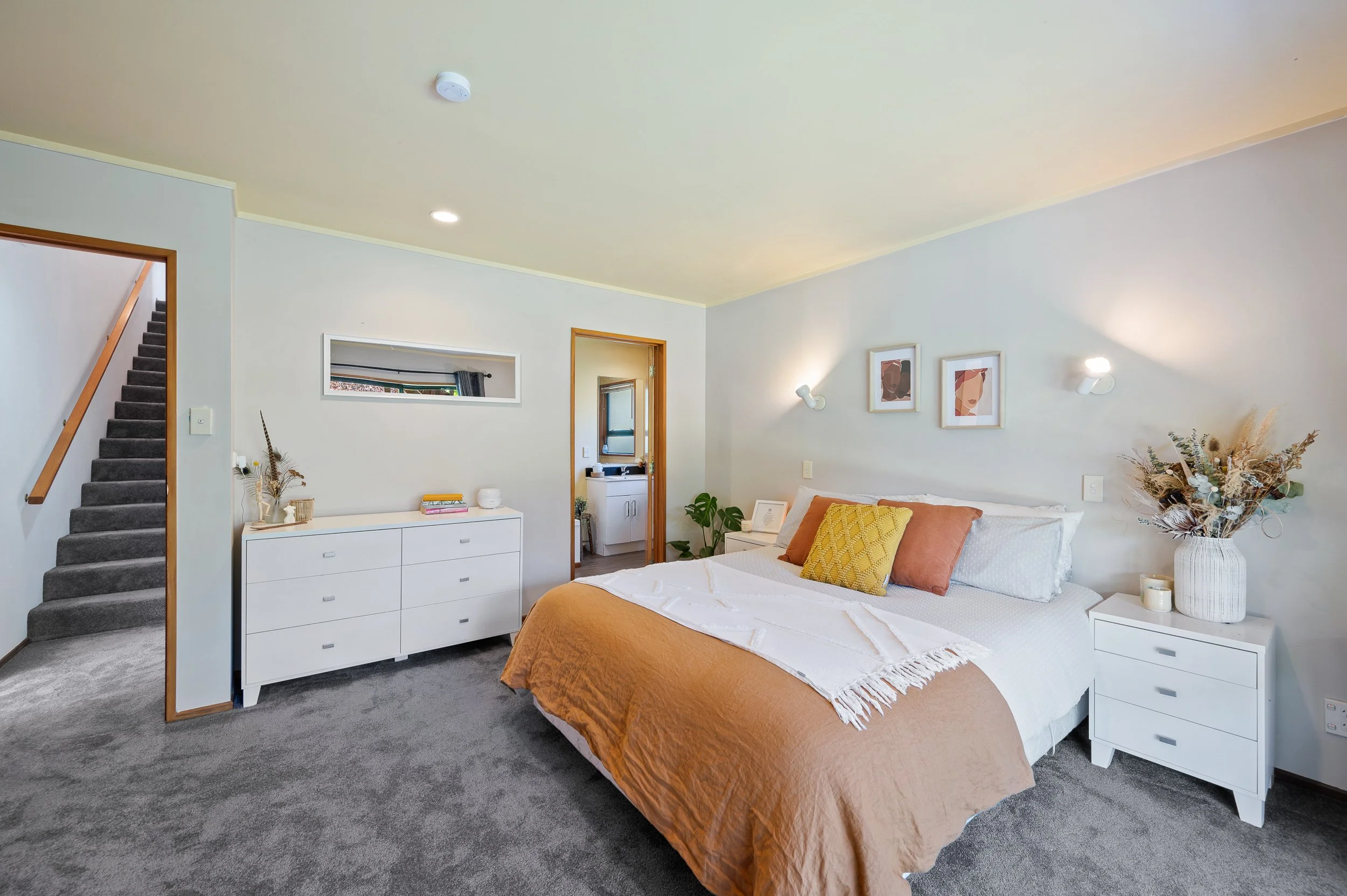 A modern bedroom with a large bed featuring white sheets, tan blankets, and pillows in yellow and rust colors. There are white nightstands on either side of the bed, one with a vase of dried flowers. A dresser with decorative items is against the wal