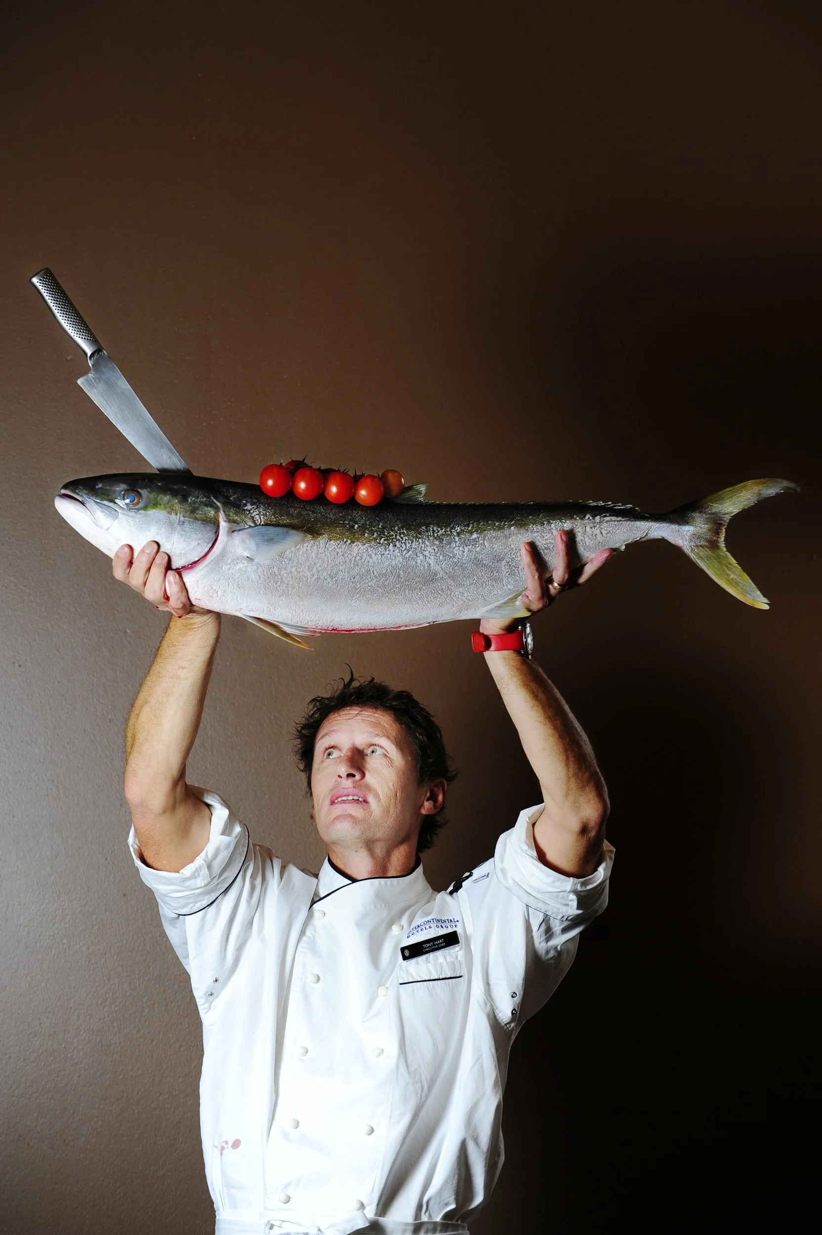 A chef holding a large fish with a bunch of cherry tomatoes on top, a knife stuck in the fish's back, against a plain brown background.