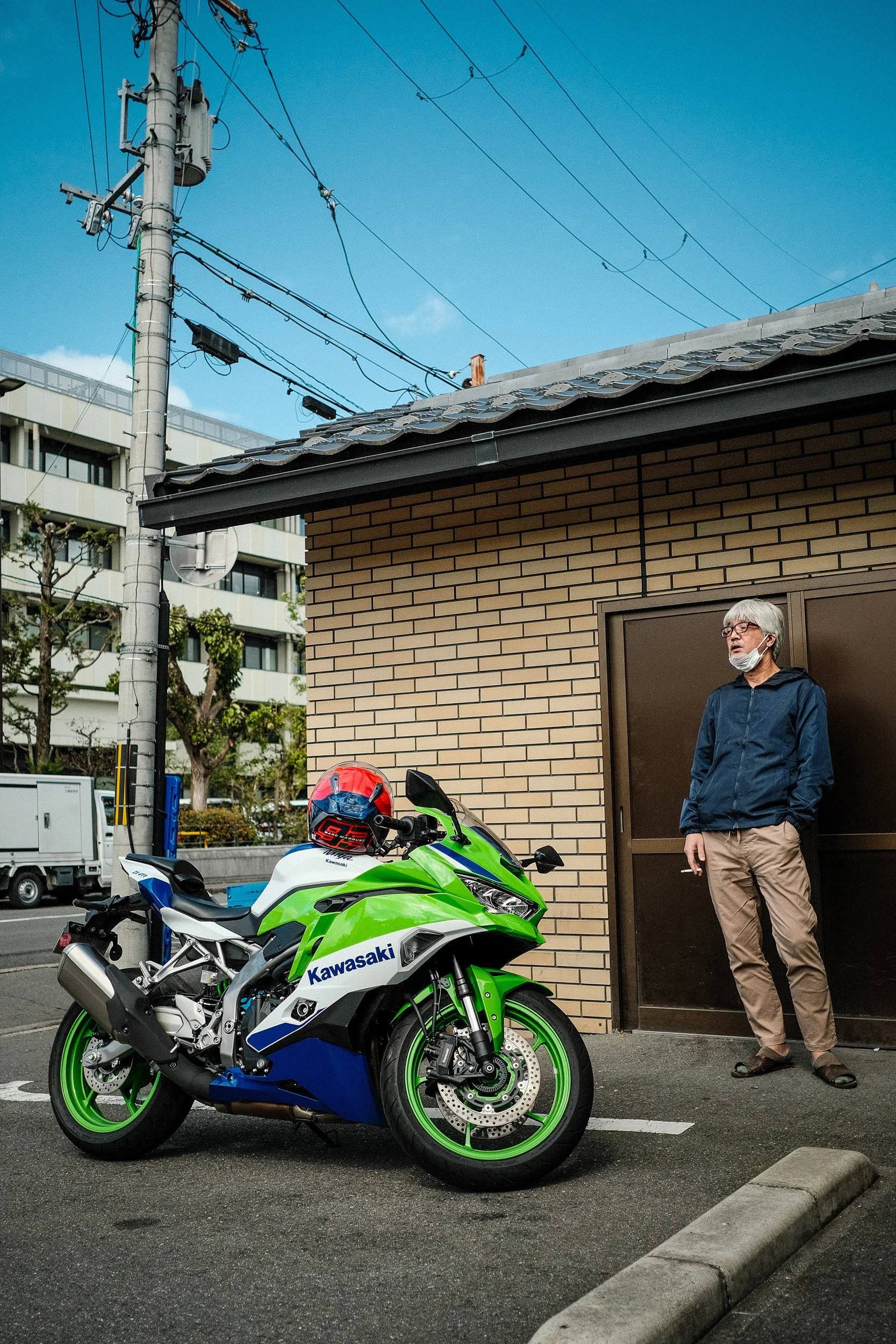 A man with gray hair, glasses, and a face mask around his neck stands beside a bright green and white Kawasaki motorcycle with a red helmet resting on the seat. The scene is outdoors with a brick building, electrical wires, and a parking lot in the b