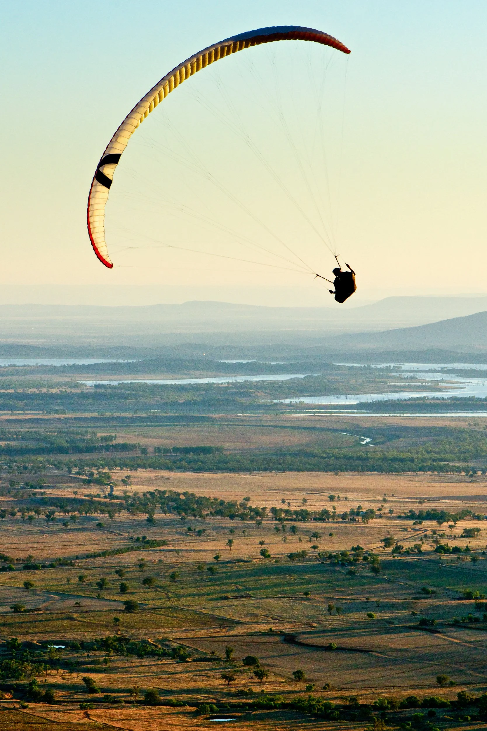 A paraglider flying high above a wide landscape with fields, rivers, and distant mountains during sunset or sunrise.