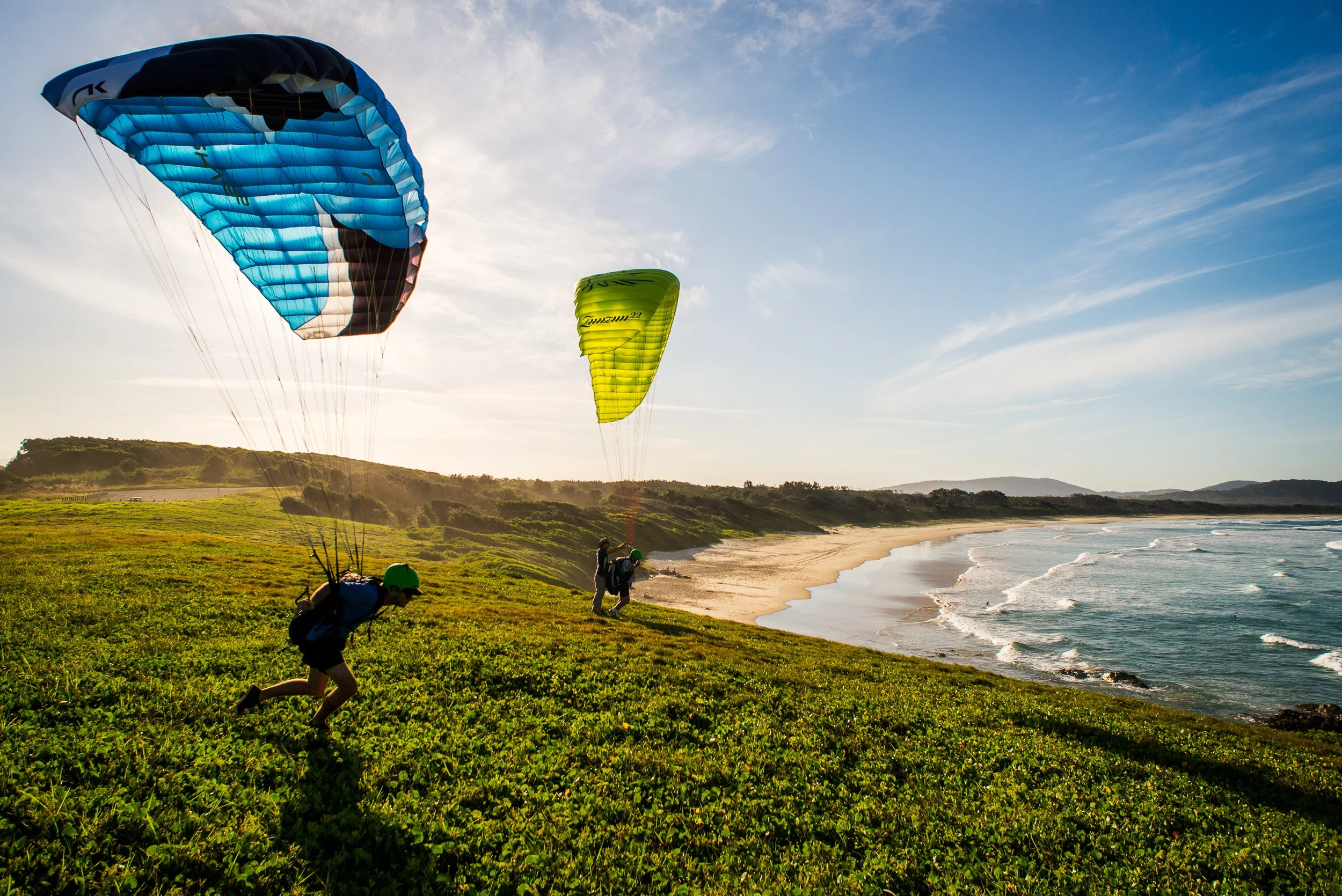 Two people preparing to land with parachutes on a grassy hill near the beach during sunset.