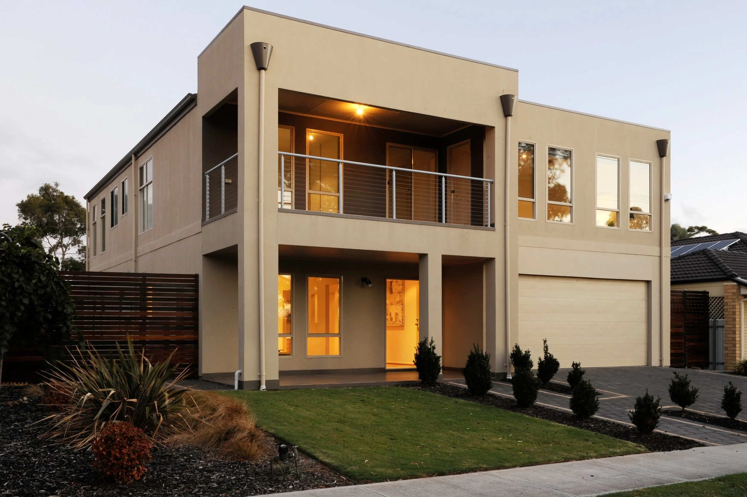 Modern two-story house with a beige exterior, large windows, a garage, and a small front yard with plants and a lawn, illuminated during twilight.