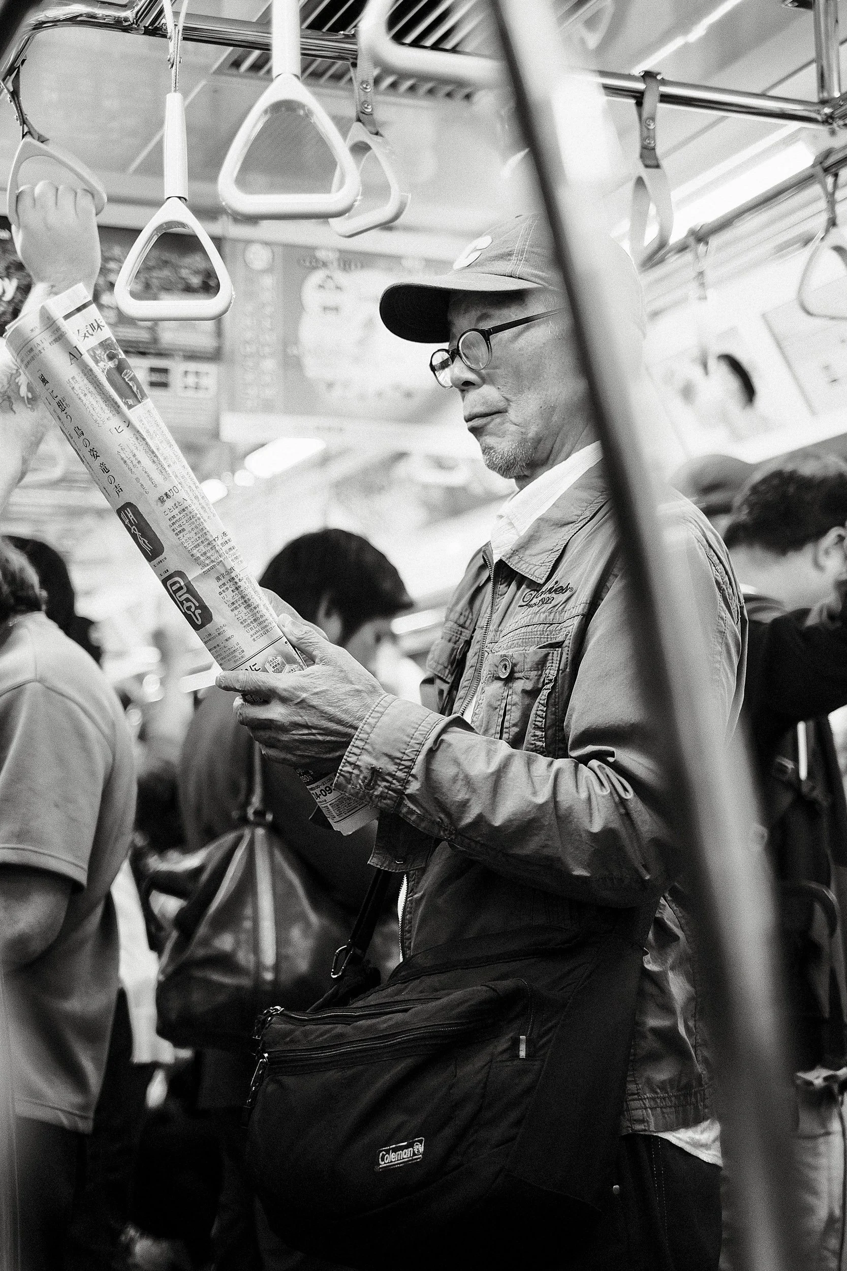 An older man with glasses and a cap stands on a crowded train, reading a newspaper.