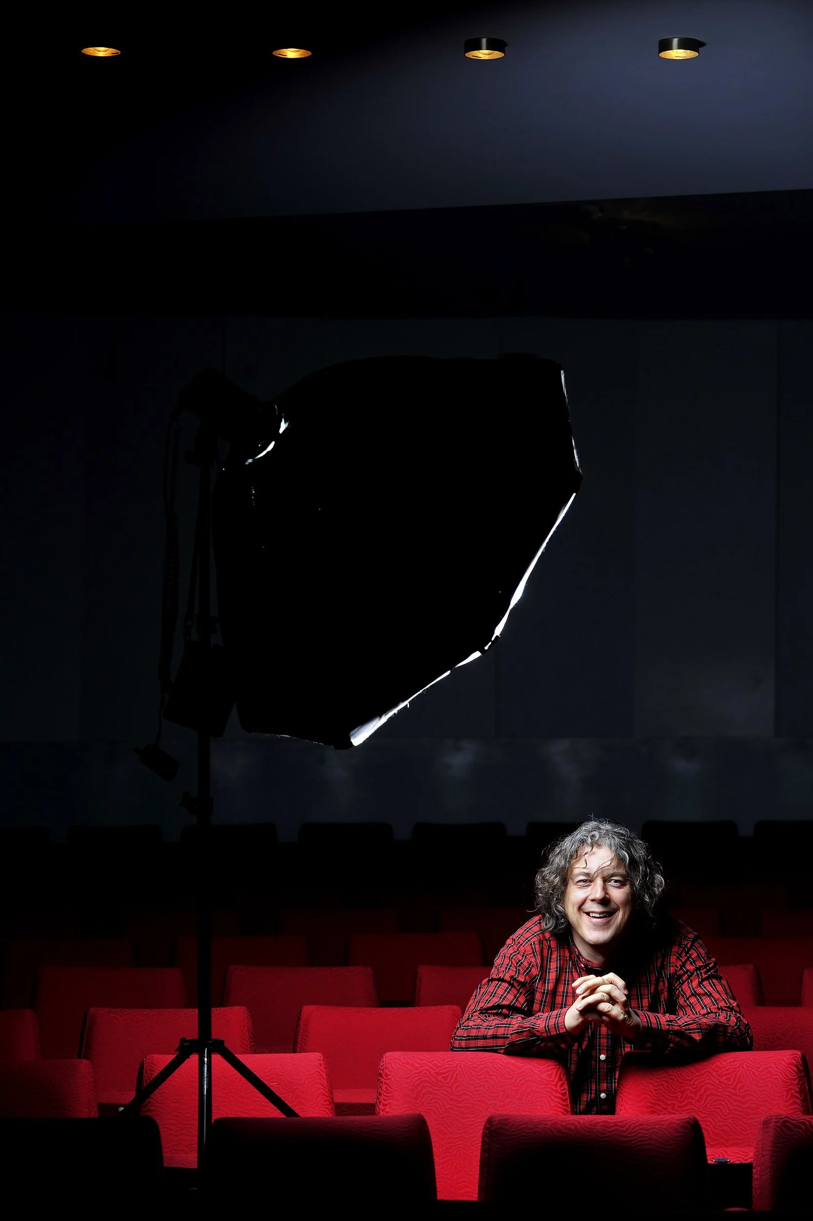 A man with curly hair and a red plaid shirt sitting in a dark theater, smiling, with studio lighting equipment overhead and empty red seats surrounding him.