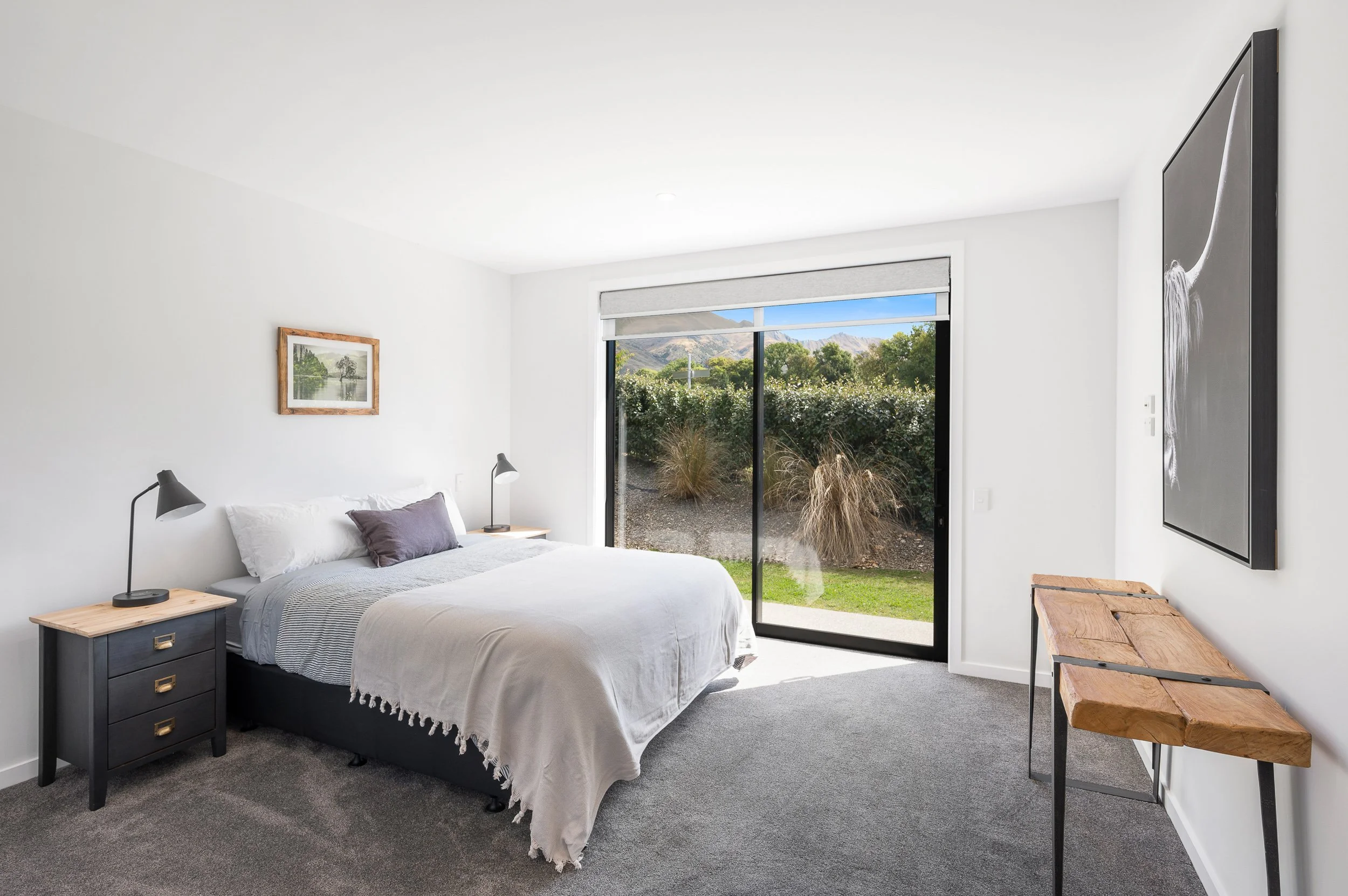 Bright bedroom with a large window showing outdoor greenery and mountains, a bed with white linens, black nightstands with lamps, a wooden console, and a wall-mounted painting.