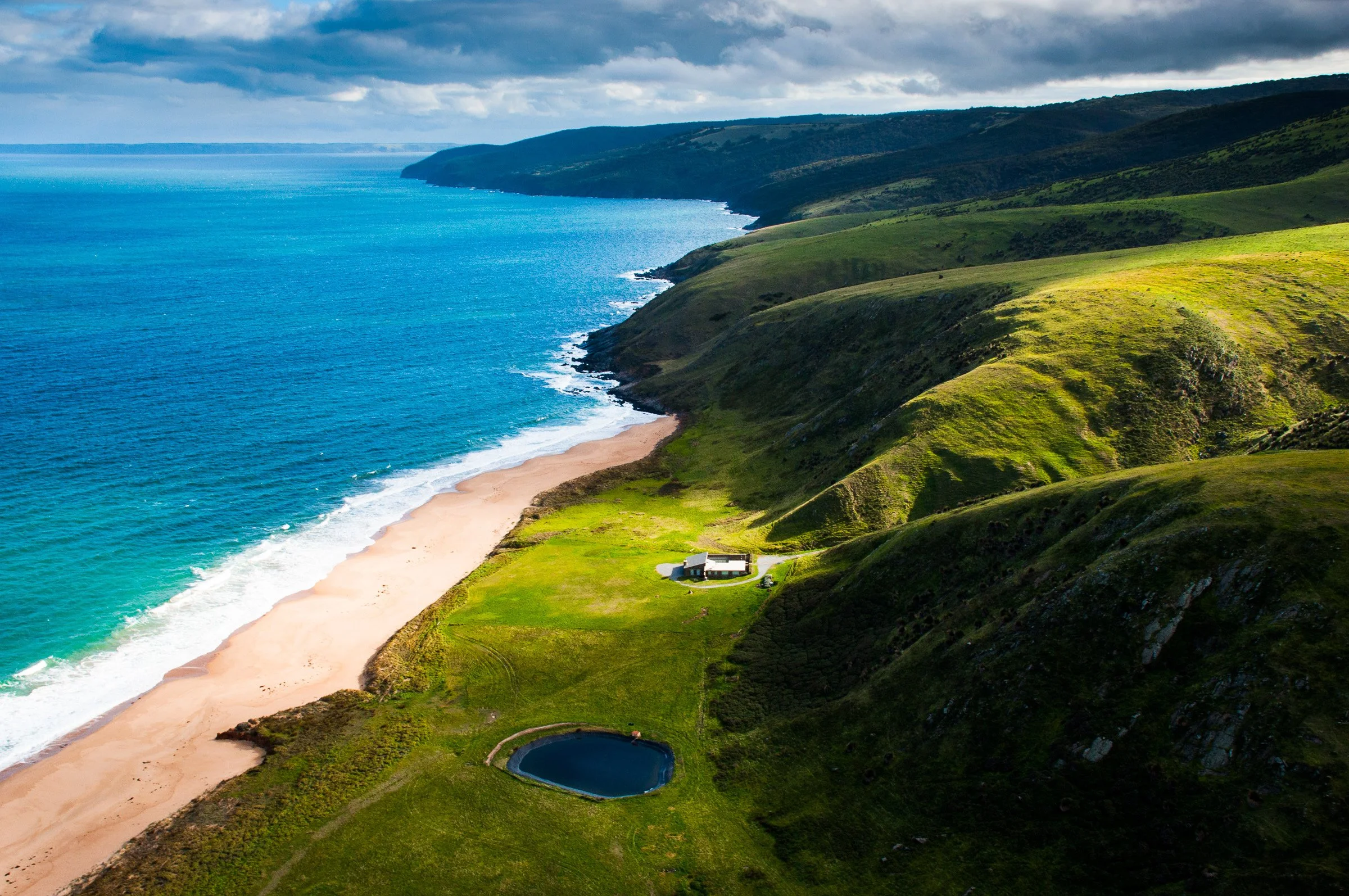 Aerial view of a coastal landscape with a sandy beach, blue ocean, green hills, and a small building near a pond.