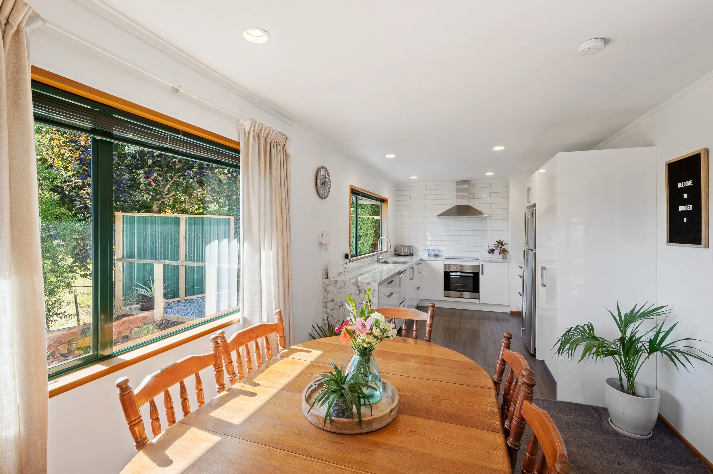 A dining area with a wooden table and chairs, a vase with flowers, large windows with curtains, and a kitchen with white cabinetry and stainless steel appliances in the background.