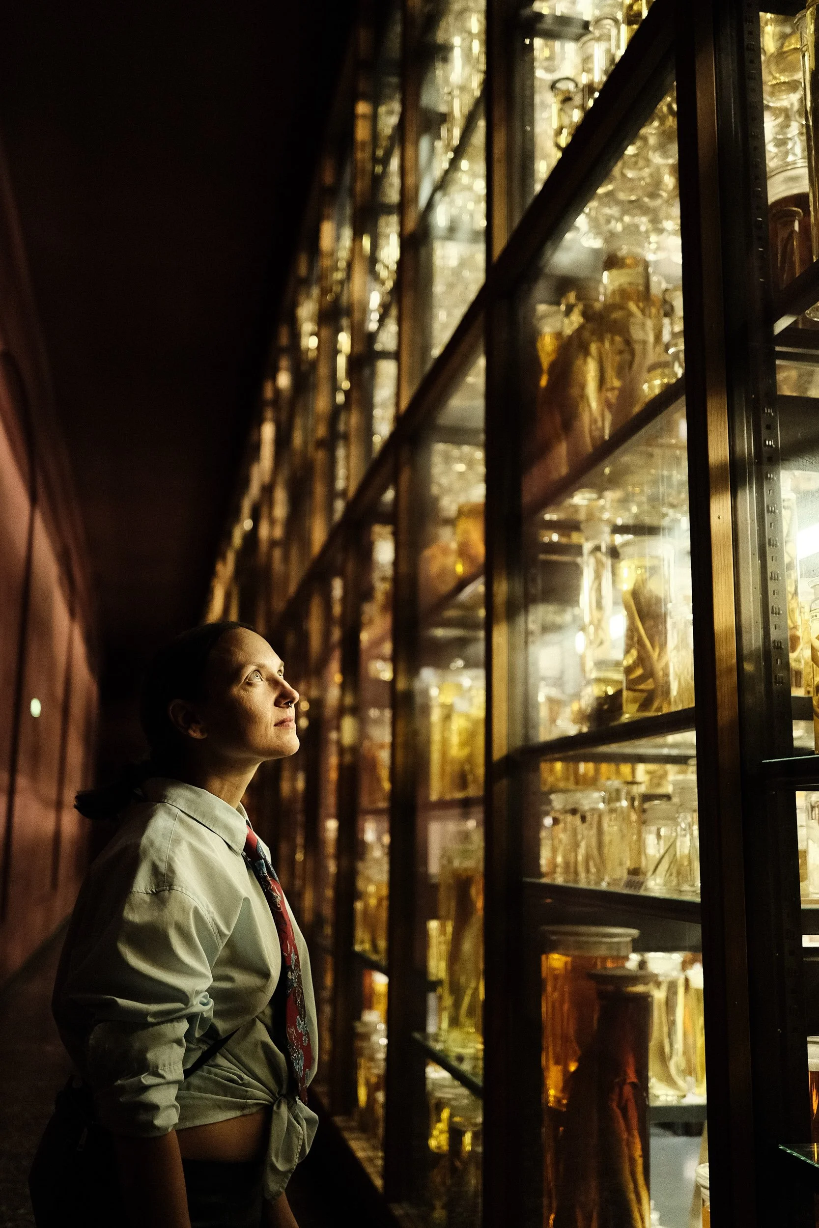 A woman in a light-colored shirt and patterned tie stands in front of glass display cases filled with preserved biological specimens illuminated from within.