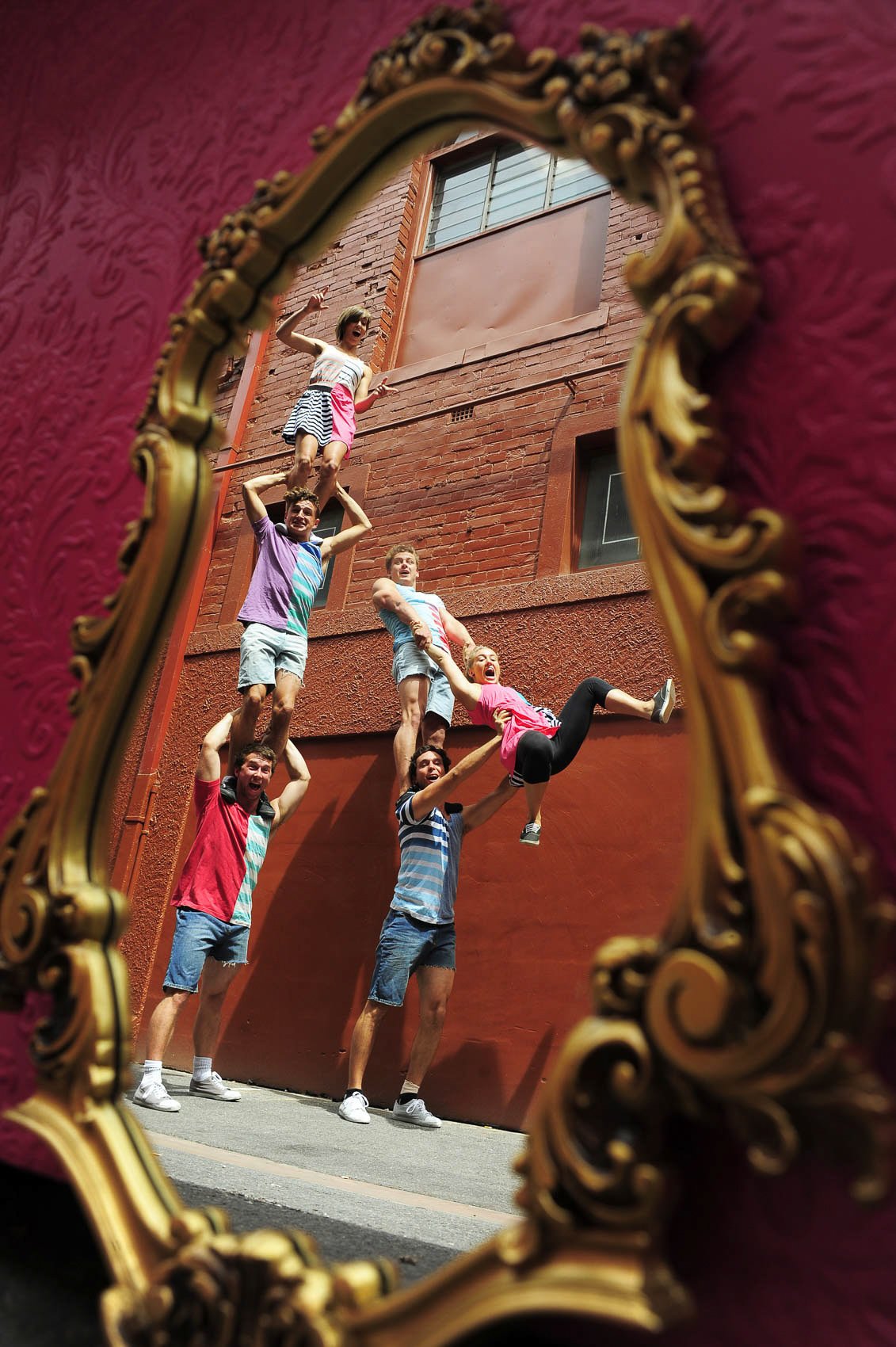 A group of seven young people forming a human pyramid, reflected in an ornate mirror, with a brick building in the background.