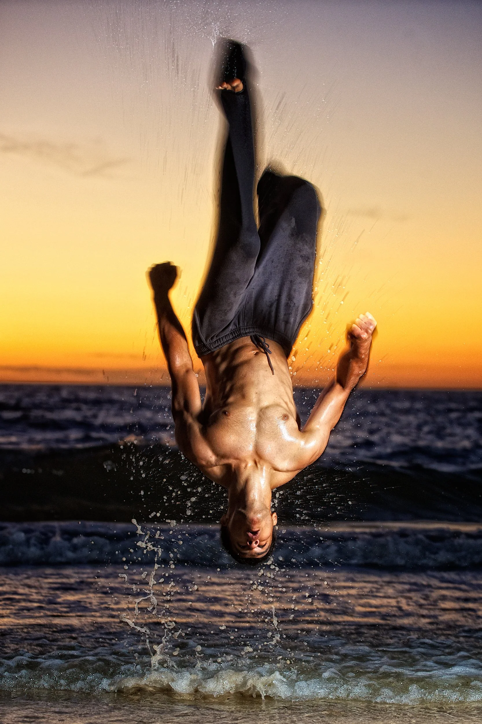 A shirtless man is mid-backflip on the beach during sunset, splashing water in the air.