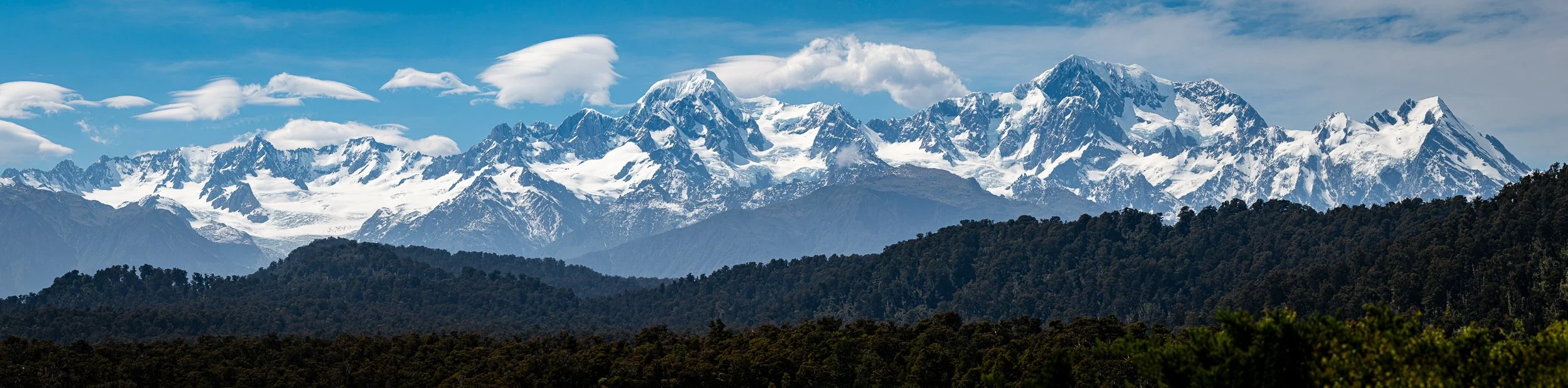 Snow-capped mountain range with a blue sky and scattered clouds over a forested landscape.