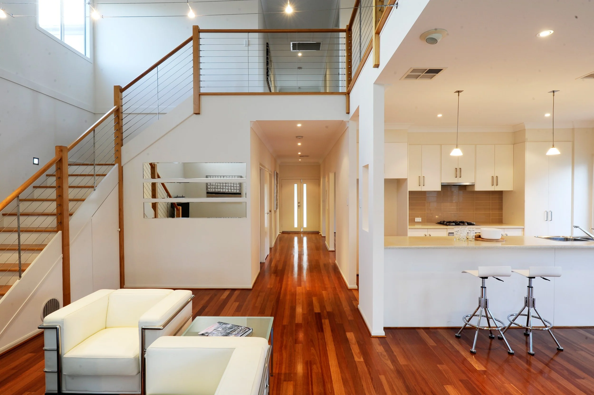 Interior of a modern, open-concept living space with a staircase, white walls, hardwood floors, a sitting area with a white couch and glass coffee table, and a kitchen with white cabinets, pendant lighting, and a breakfast bar with two stools.