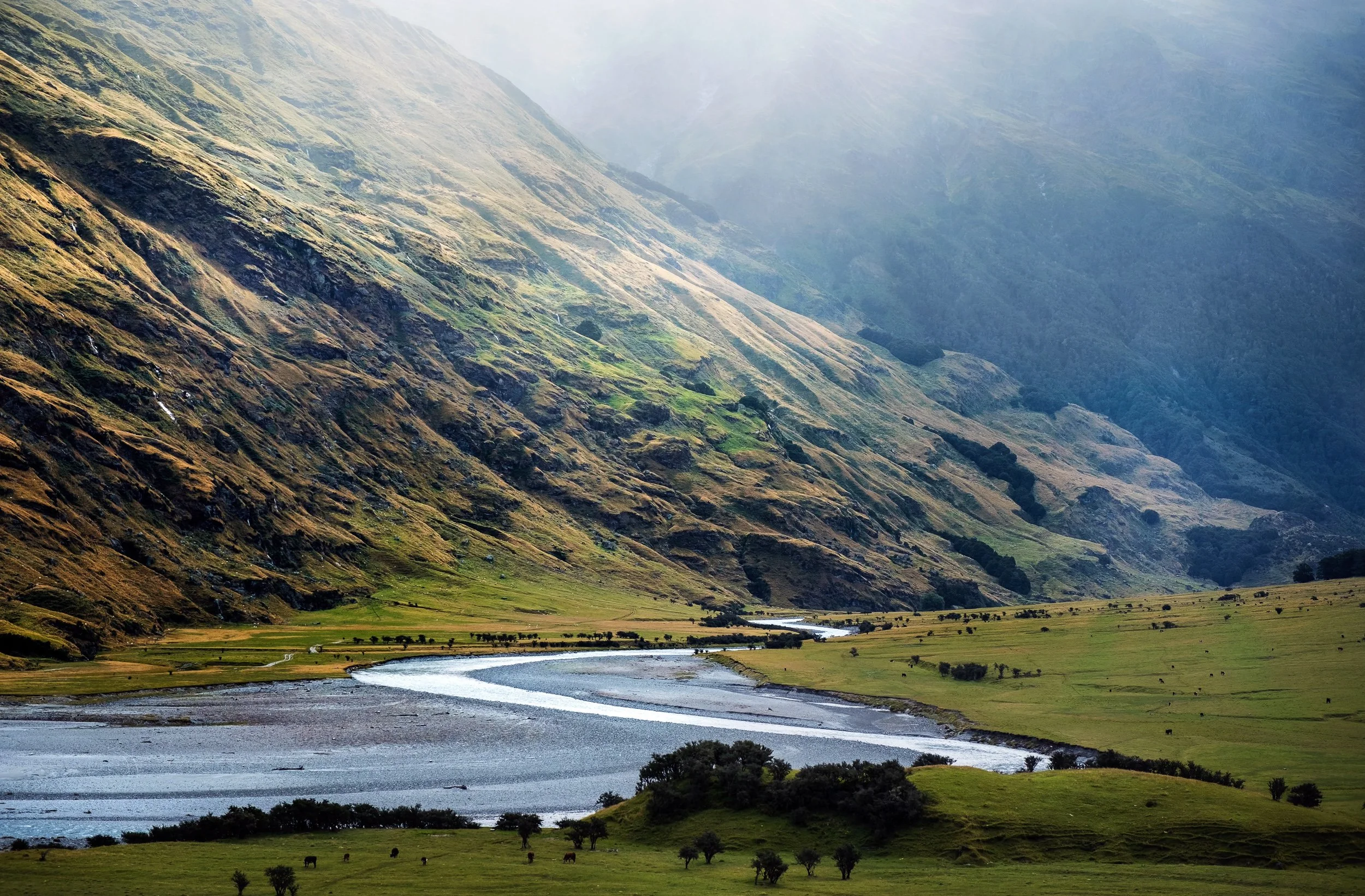 A scenic view of a valley with a river running through lush green fields, surrounded by tall, steep mountains with patches of green vegetation and some trees, under a hazy sky.