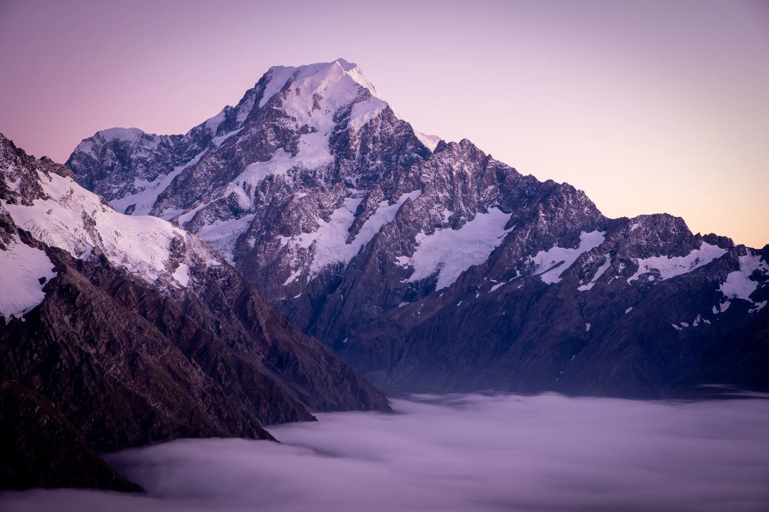 Snow-covered mountain peak with rugged cliffs, purple sky, and clouds in the valley