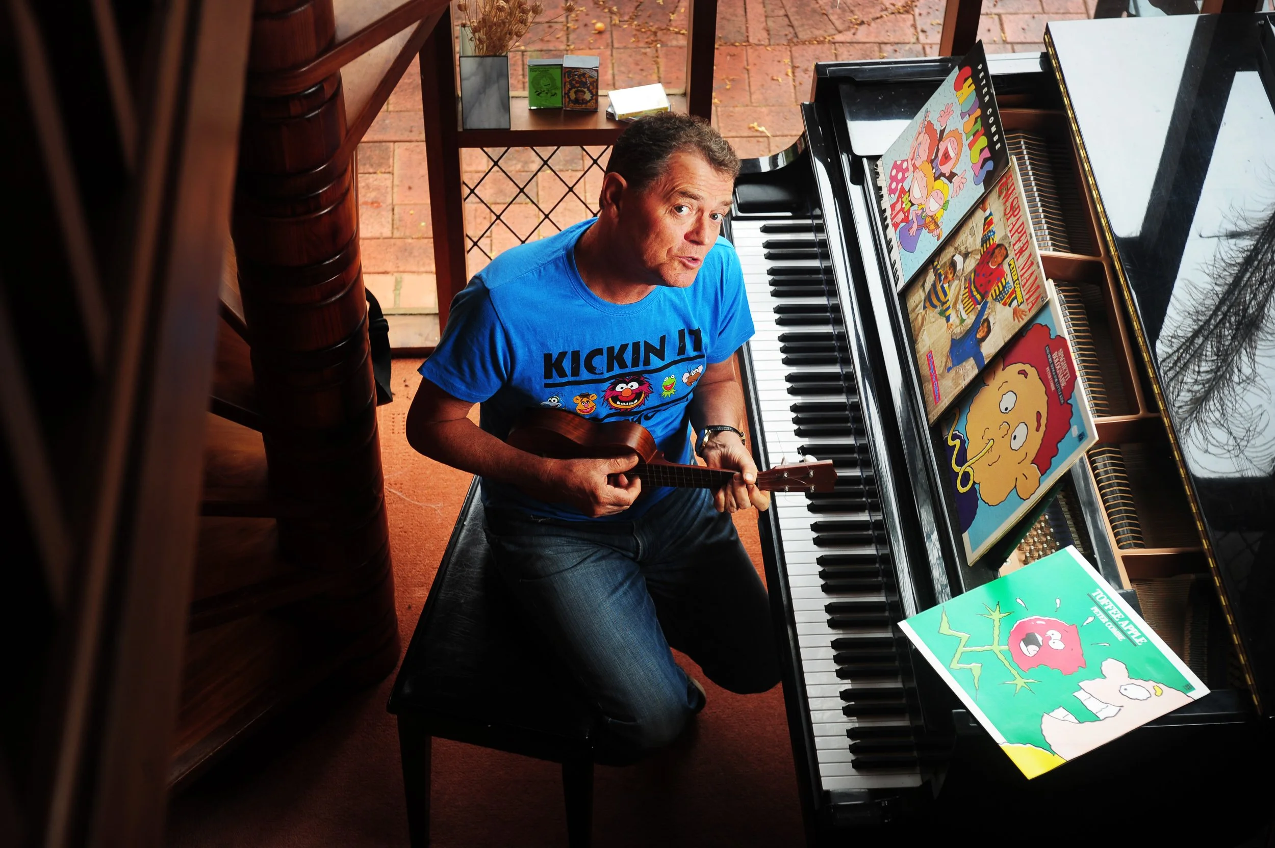 A man in a blue T-shirt sitting on a piano bench, holding a ukulele, sitting at a black grand piano with cartoon books and a magazine on top, inside a room with brick flooring and a wooden staircase.