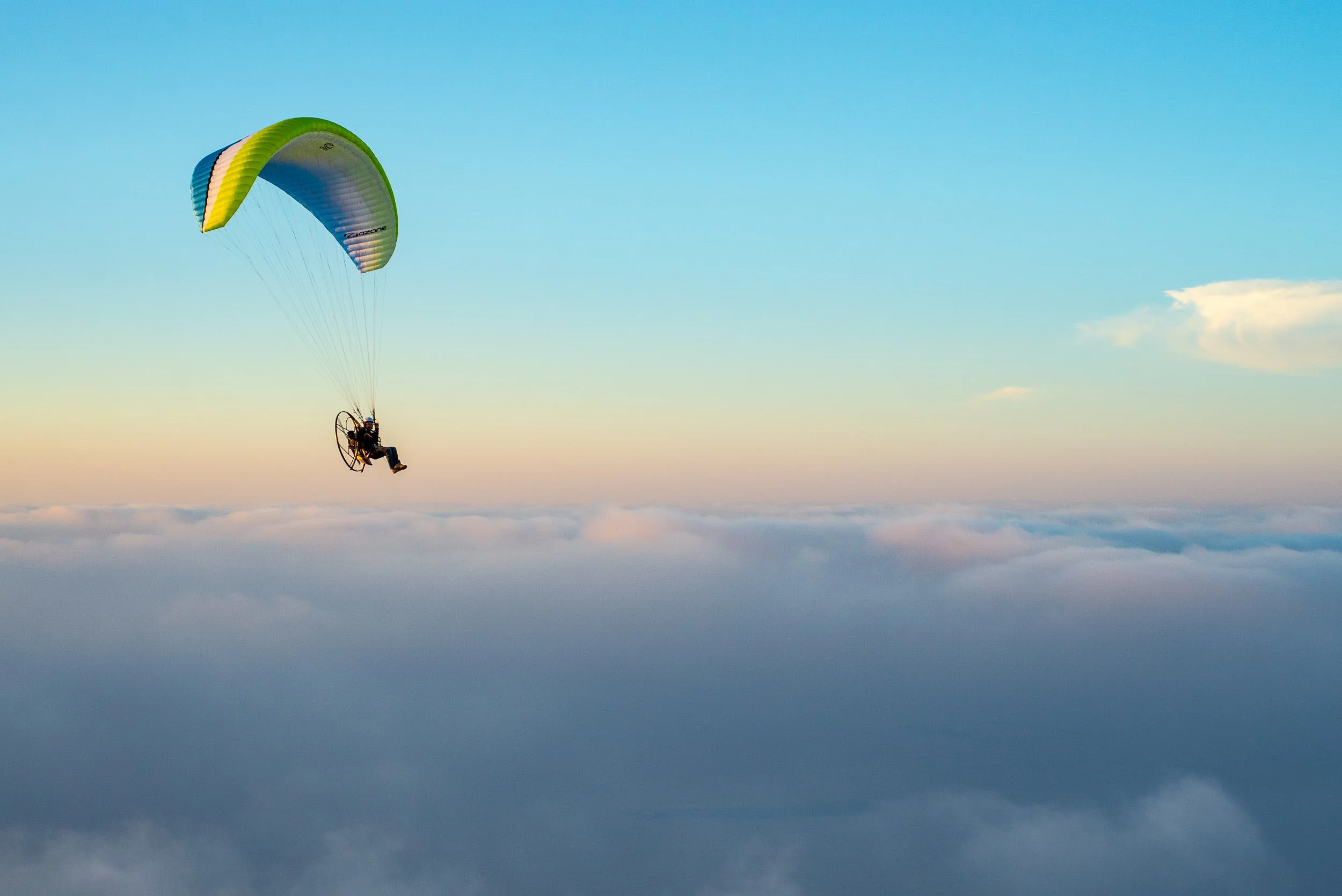 A person flying a powered parachute high above the clouds during sunset or sunrise.