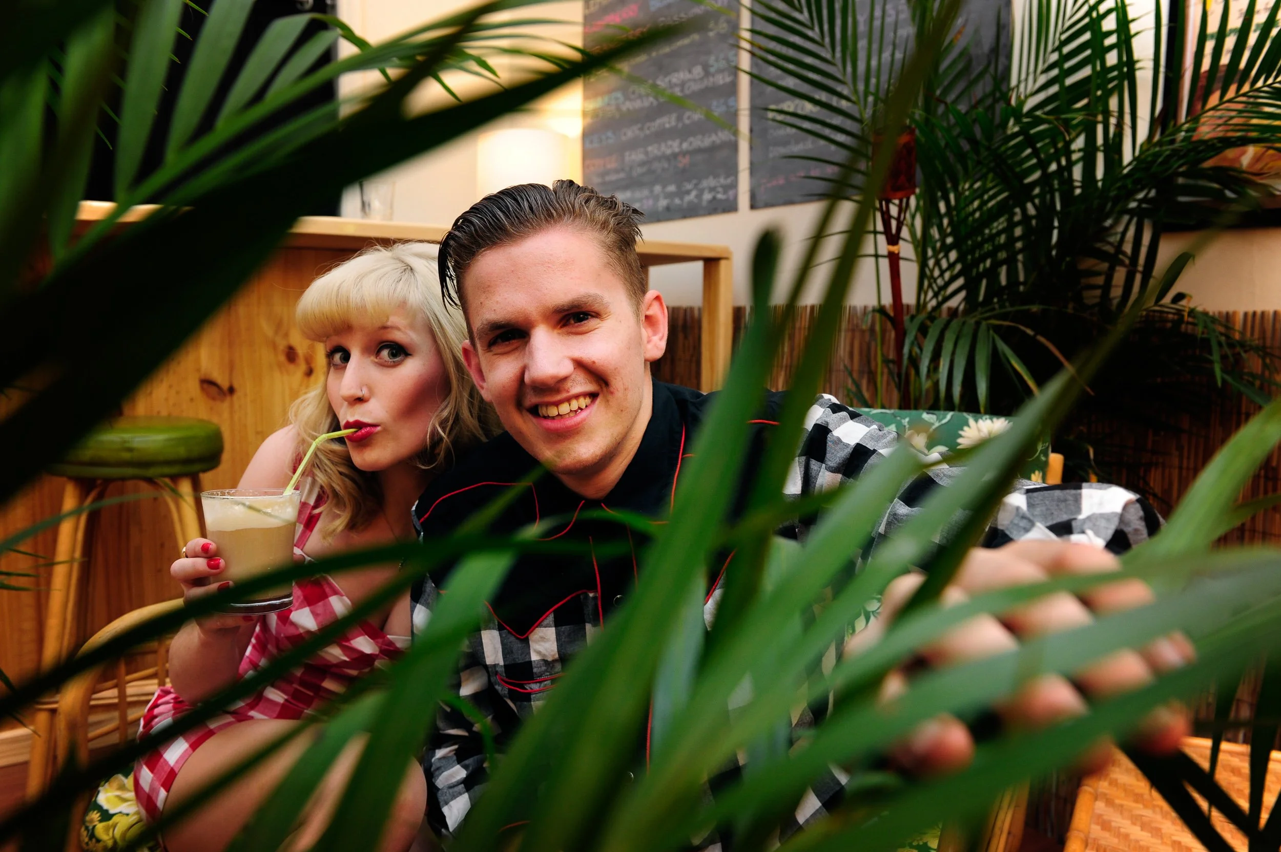 Two young adults, a blonde woman and a man with dark hair, sitting in a cozy cafe surrounded by green plants. The woman drinks a latte with a straw, while the man smiles at the camera. The cafe has wooden decor and menu boards in the background.