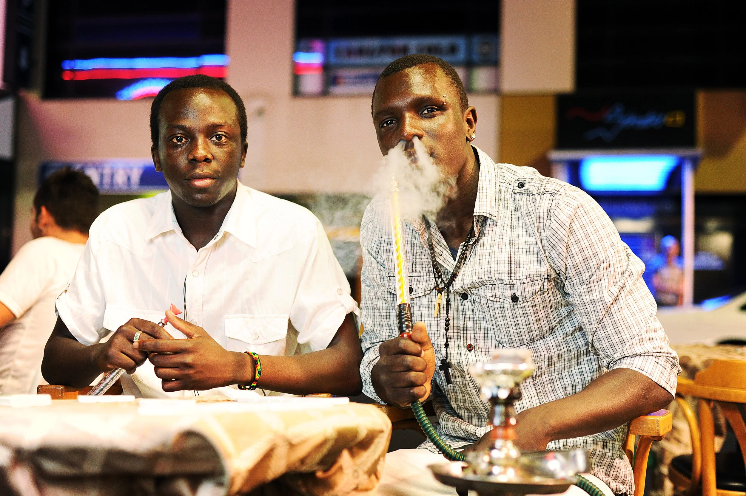 Two young men sitting in a restaurant, one of them smoking a hookah with a cloud of smoke, while the other looks directly at the camera. The background includes a screen displaying neon lights and a blurred figure.